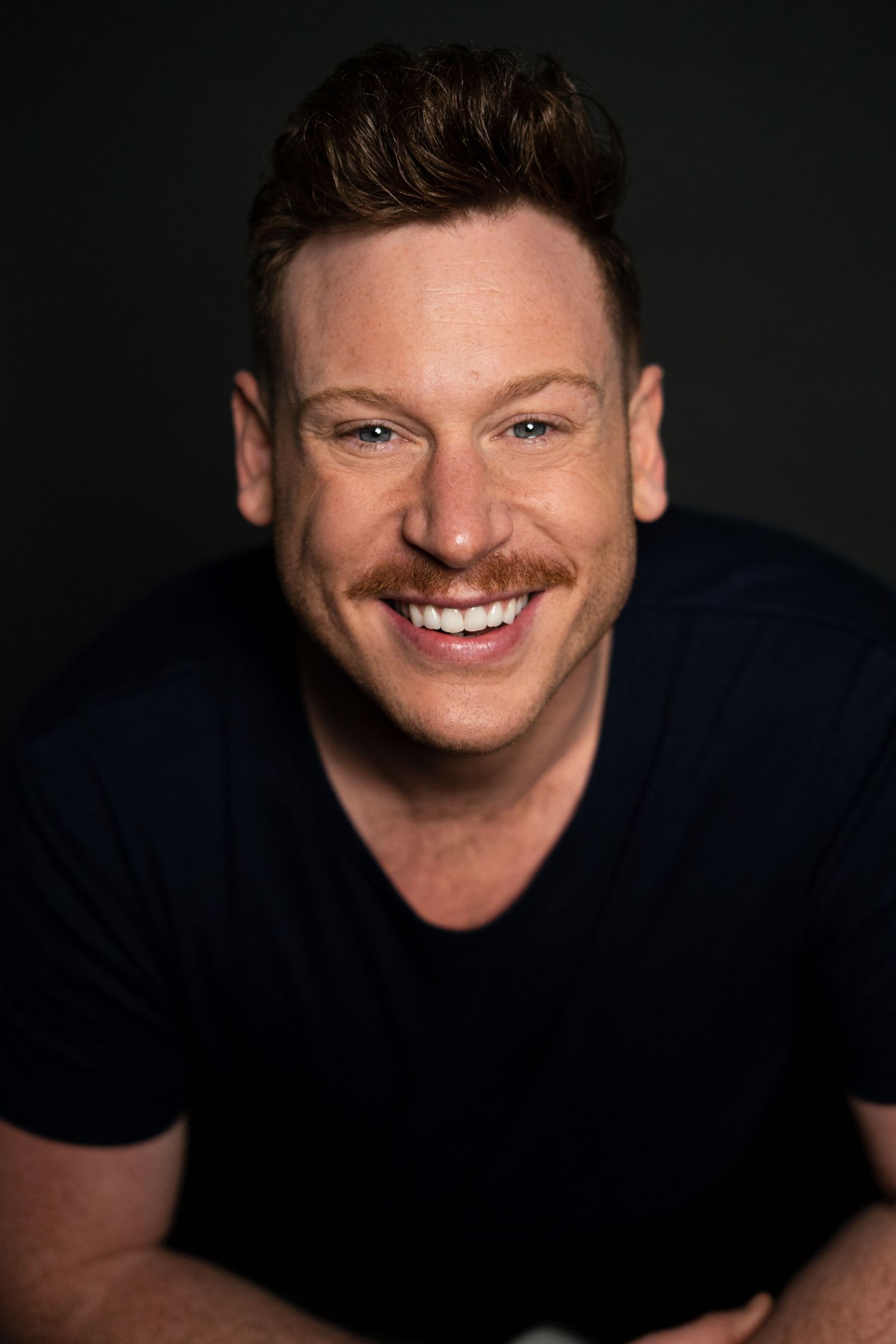 Cheerful man with reddish-brown hair and mustache in a navy t-shirt, smiling broadly against dark backdrop.