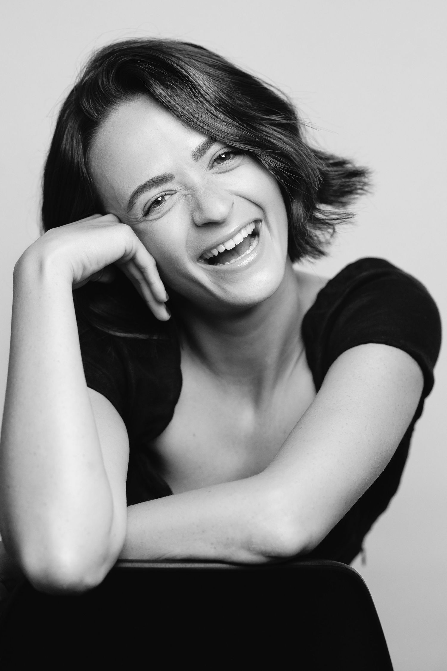 Black and white photo of a young woman with short hair laughing joyfully, resting chin on hand.