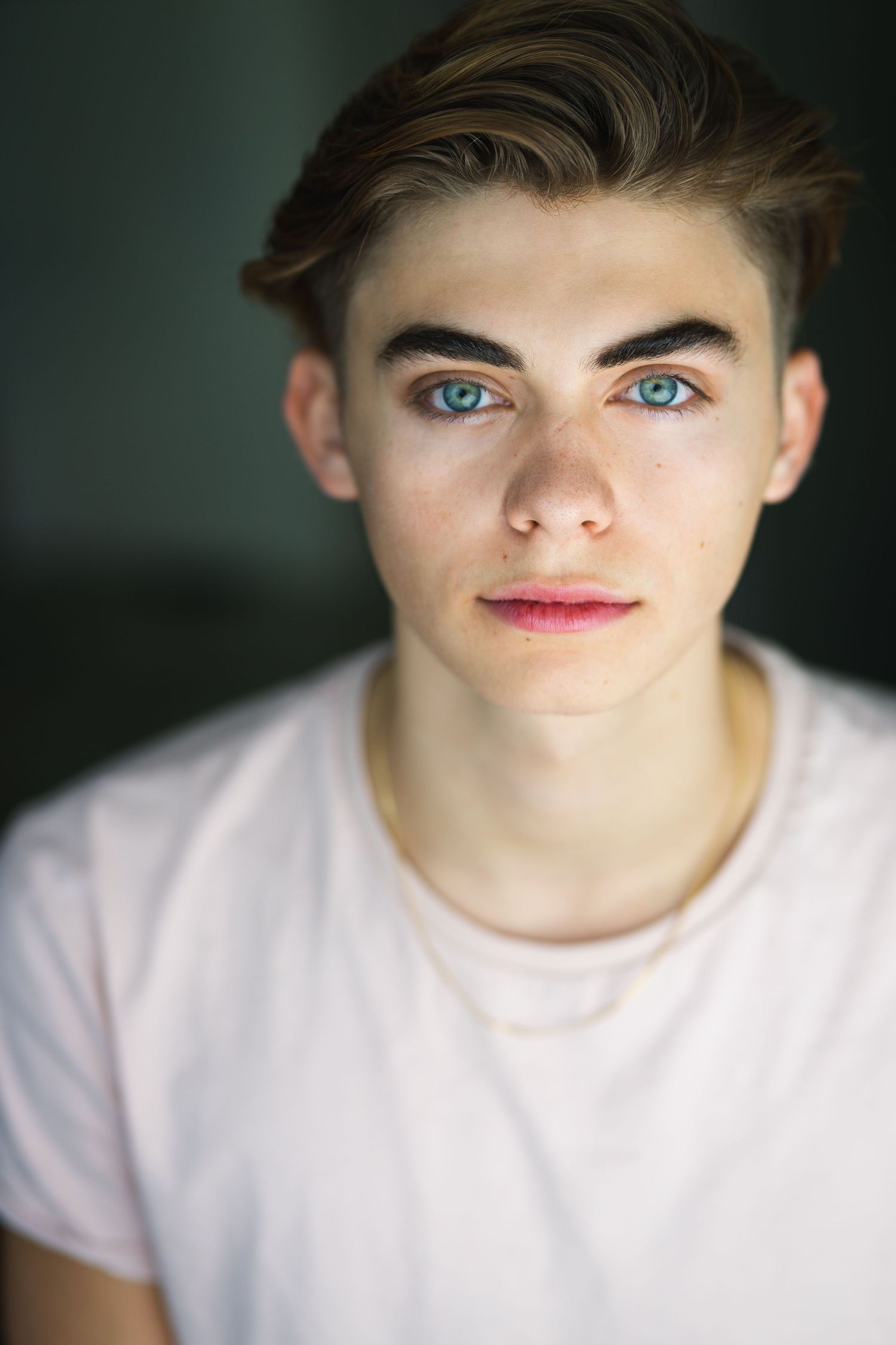 Young man with striking blue eyes and brown hair wearing a light pink t-shirt poses for a close-up portrait photo.