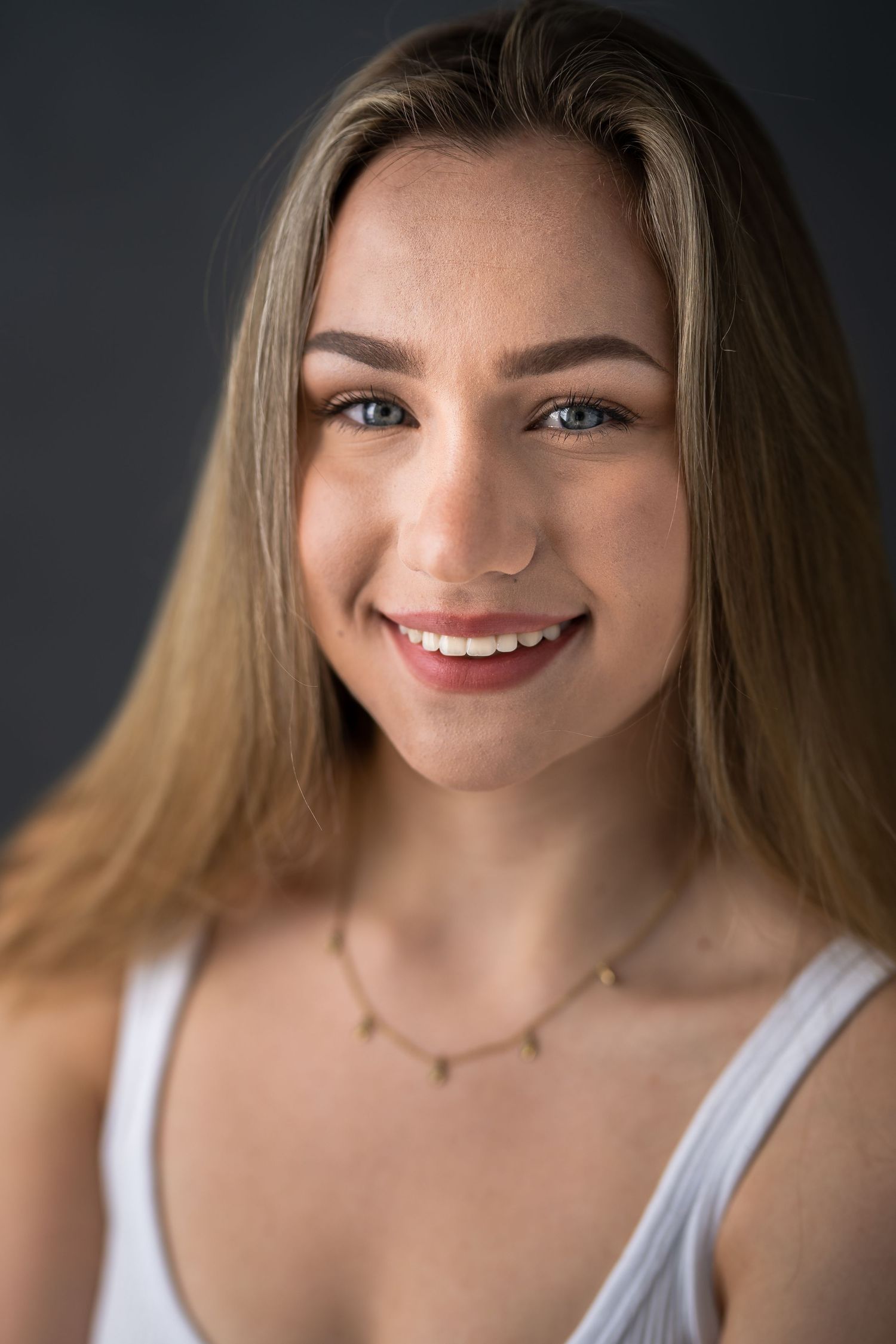 Smiling young woman with long blonde hair, blue eyes, and a gold necklace wearing a white top against gray background.
