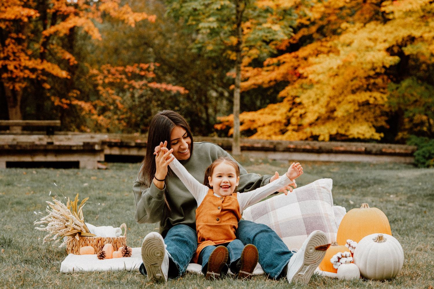 Mother and toddler enjoy a fall outdoor photo session with pumpkins and autumn foliage in the background.