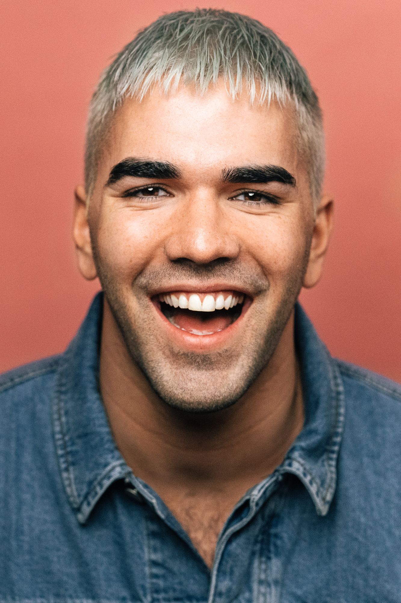 Laughing man with bleached blonde hair and dark eyebrows wearing a denim shirt against a coral background.