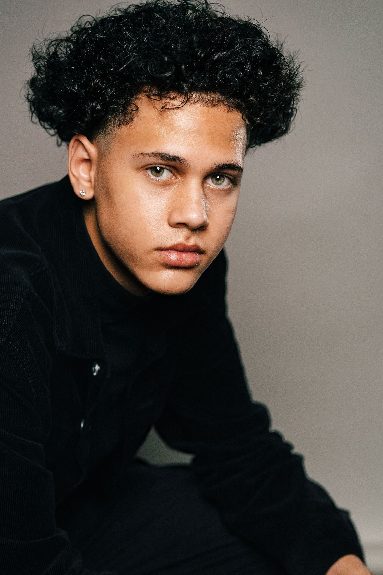 Young man with curly black hair and green eyes wearing a black outfit, posing against a gray background.