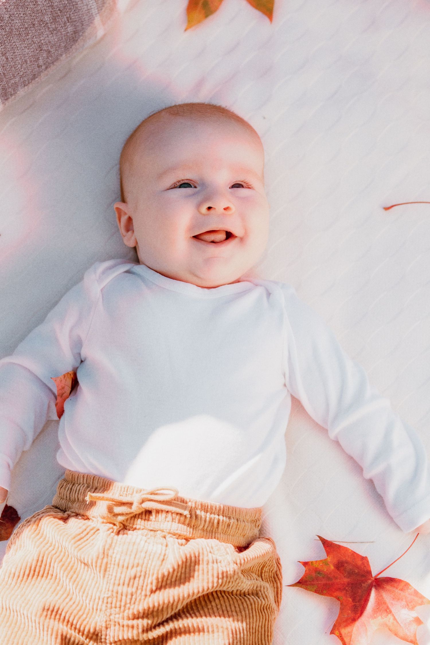 Smiling baby in white onesie and tan pants lying on white blanket surrounded by autumn leaves.