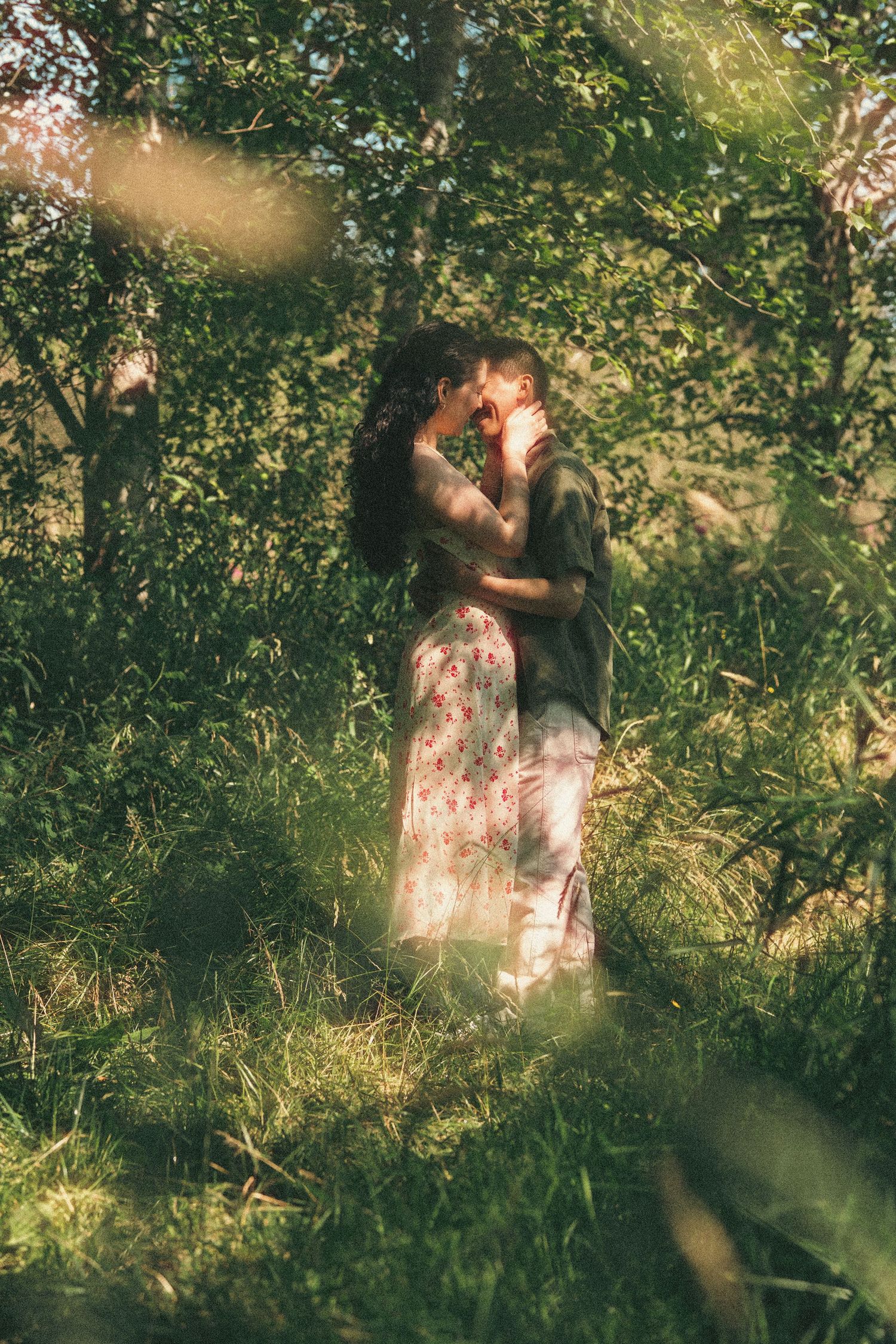 A couple embraces tenderly among lush green trees in golden sunlight, sharing an intimate moment in nature.