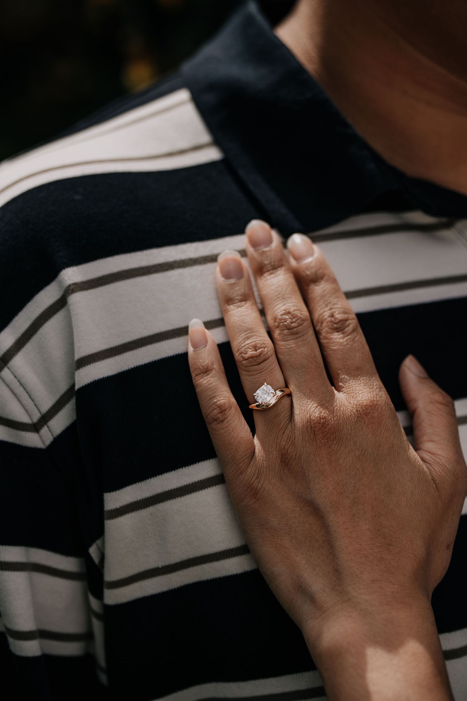 Close-up of a hand wearing a stunning diamond engagement ring, resting on a striped navy and white shirt.