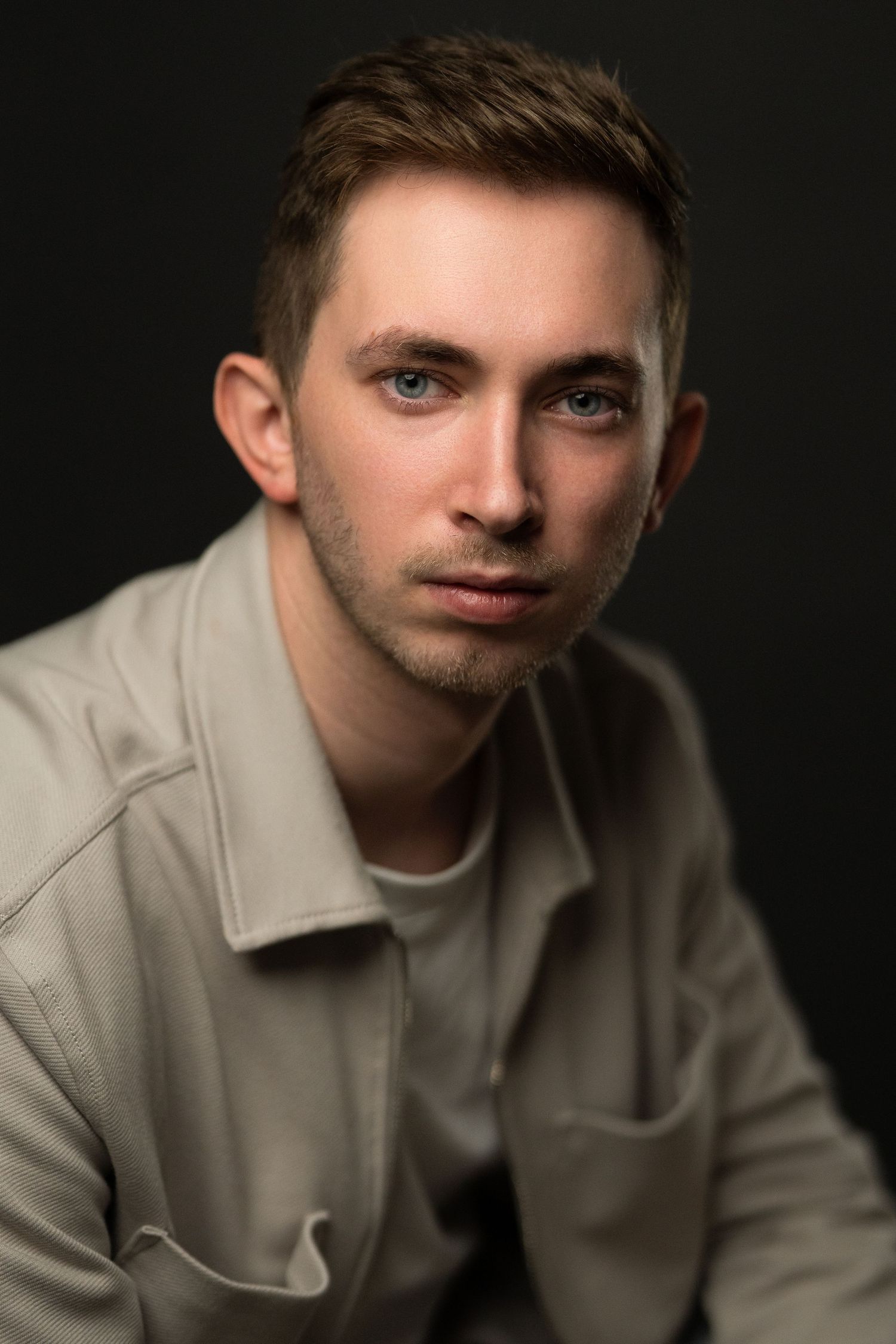 Young man with blue eyes and light brown hair wearing a beige jacket, posed against a dark background.