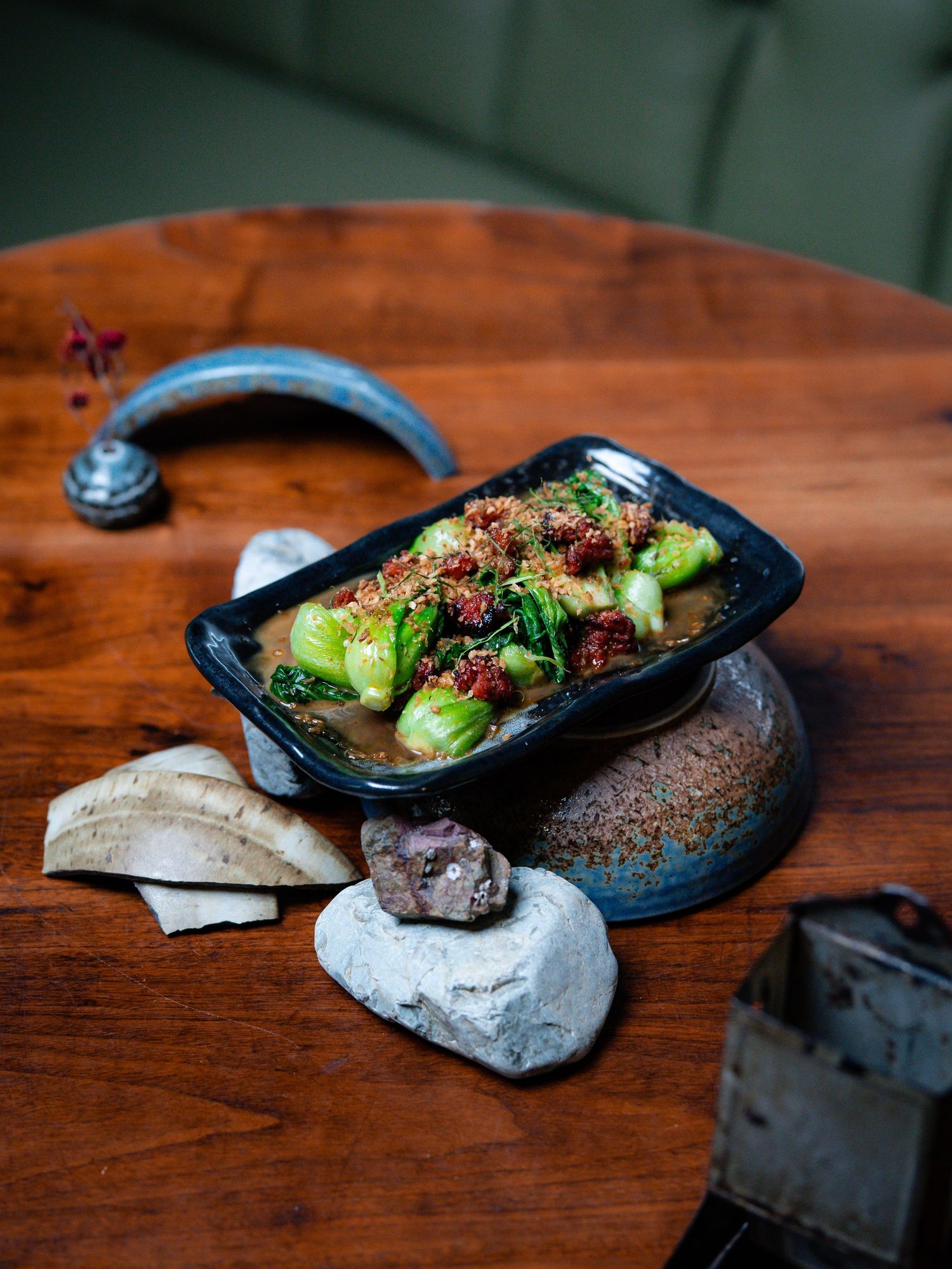 Black ceramic dish with braised bok choy, spiced minced meat, and sesame seeds, served on a wooden table with decorative stones.