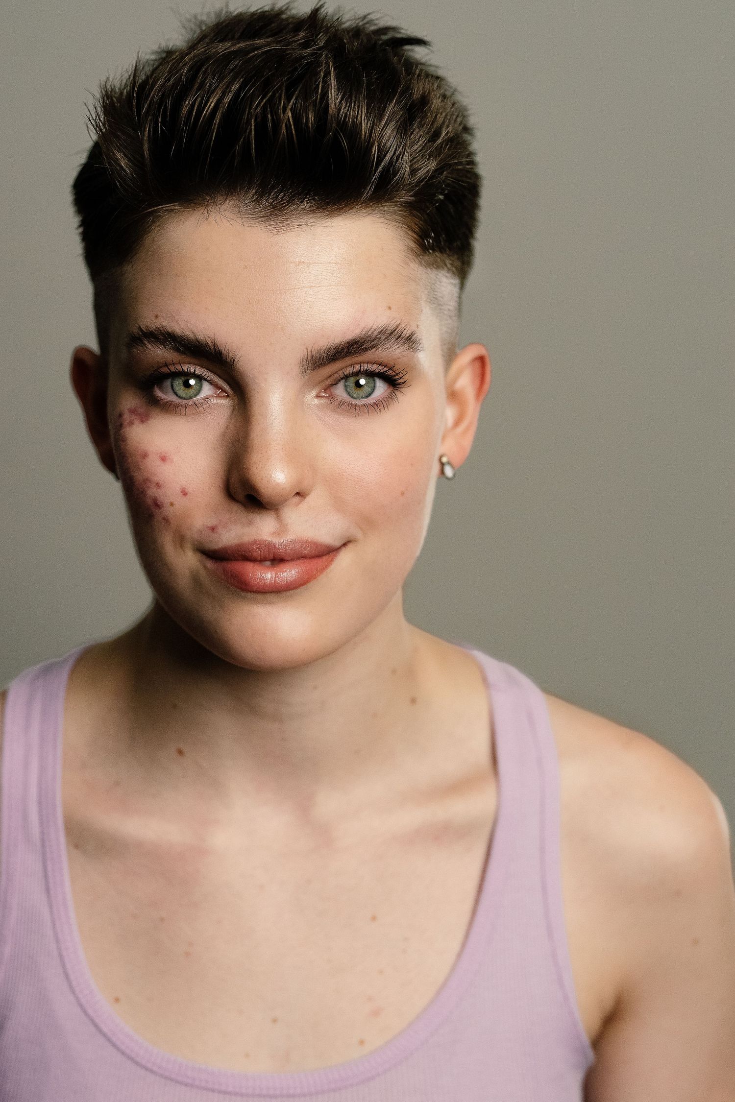 Young person with short dark hair, green eyes, and a lavender tank top poses for a studio portrait.