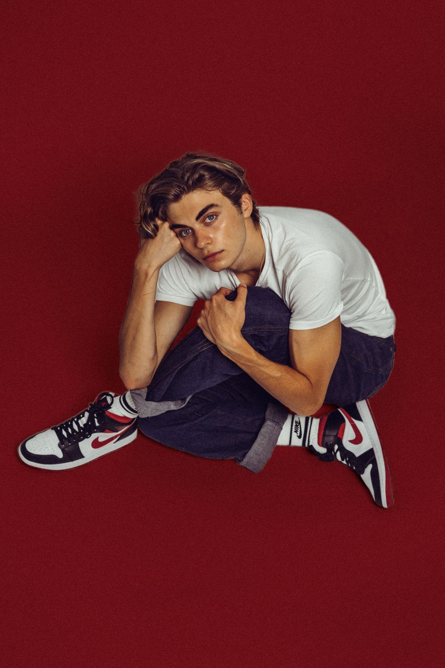 Young man in white tee, jeans, and Nike Air Jordan 1s sits on red background in a stylish editorial pose.