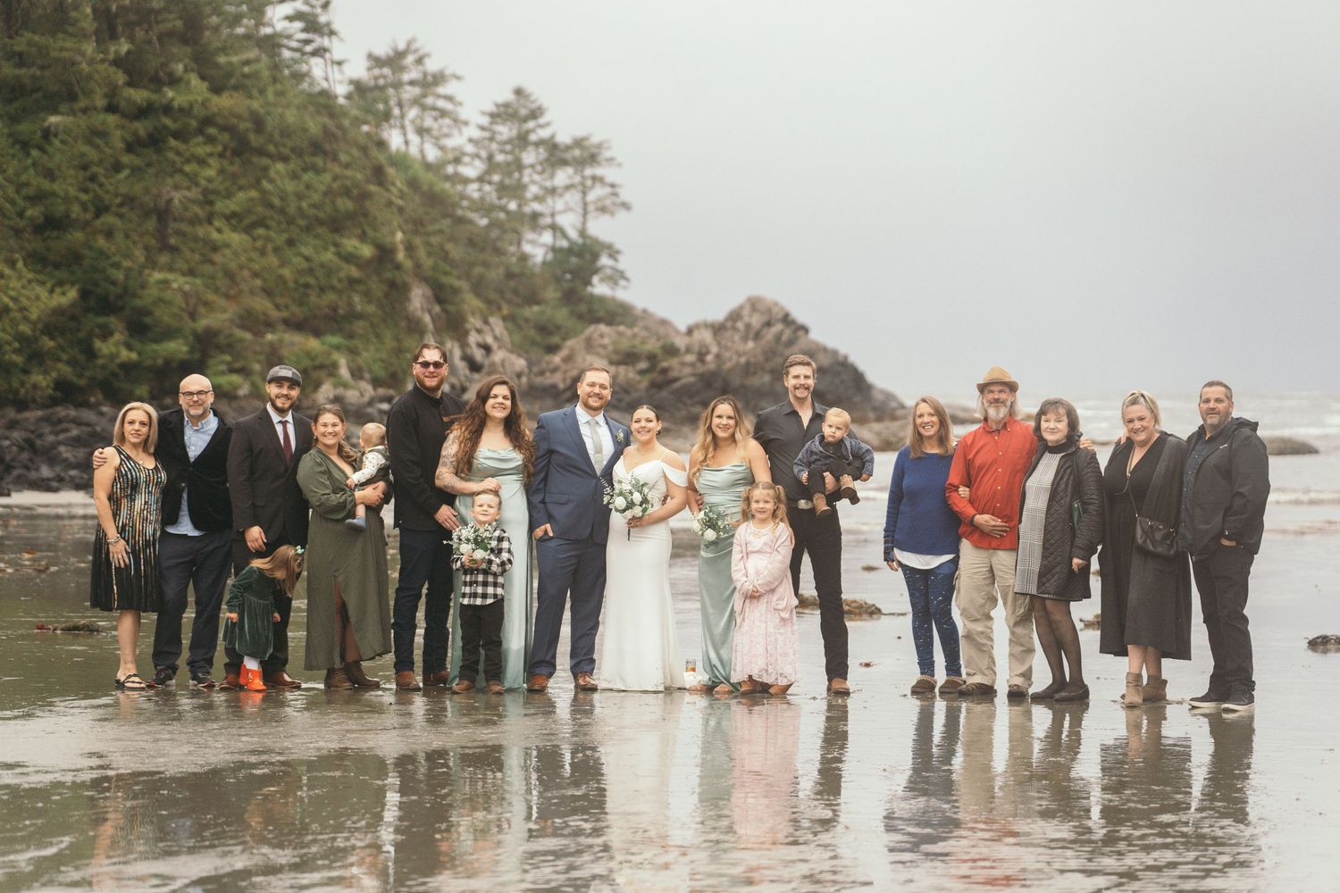 Wedding party poses on a misty beach with rocky coastline and evergreen trees in the background.