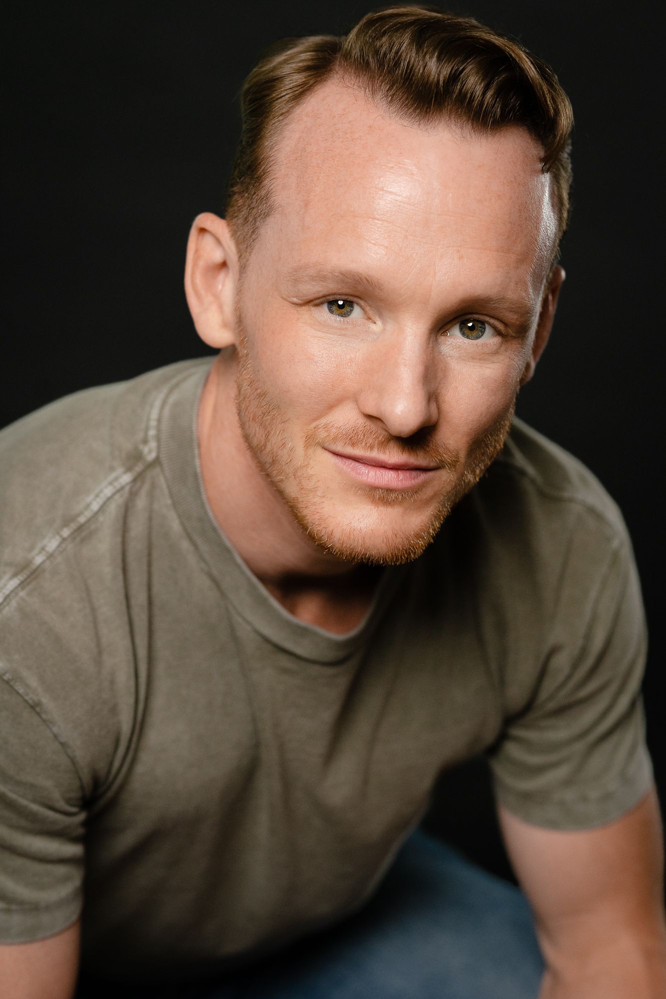 A smiling red-haired man in an olive green t-shirt poses against a dark background in a professional headshot.
