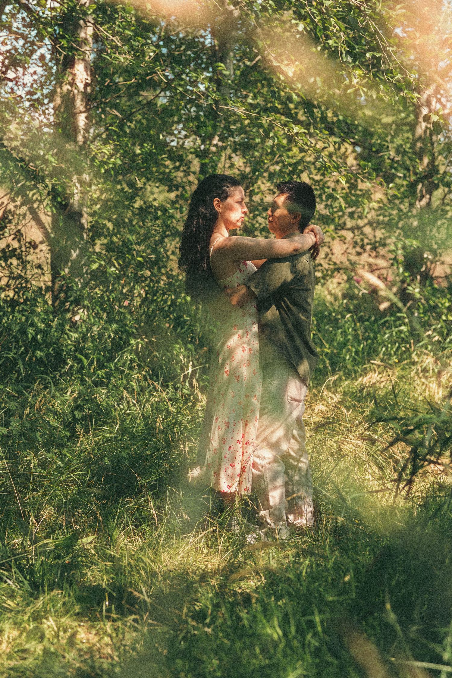 A couple embraces romantically in a sunlit green forest, the woman in a floral dress and man in dark attire.