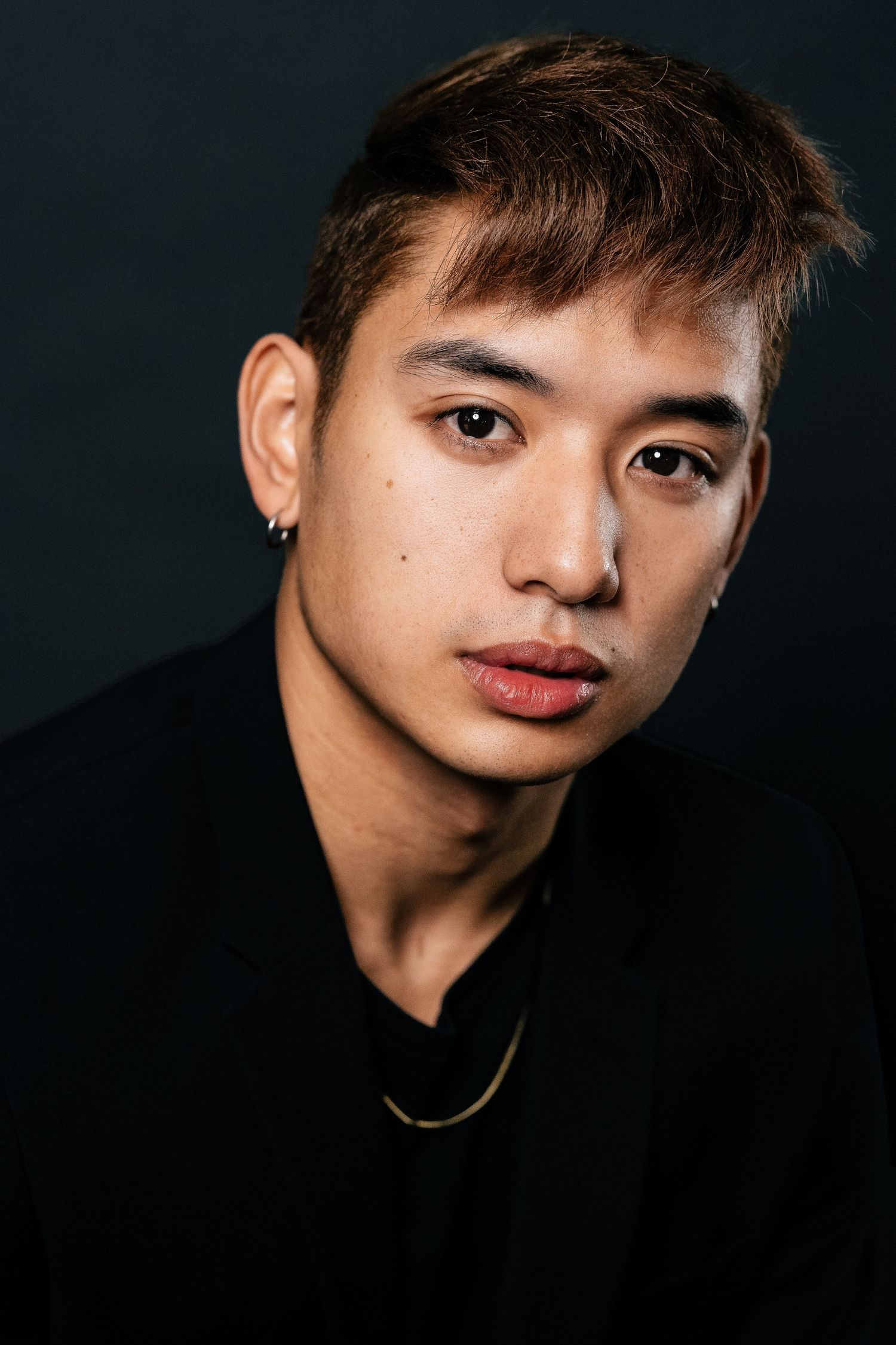 Young Asian man with brown hair and small earring wearing black shirt and gold necklace against dark background.