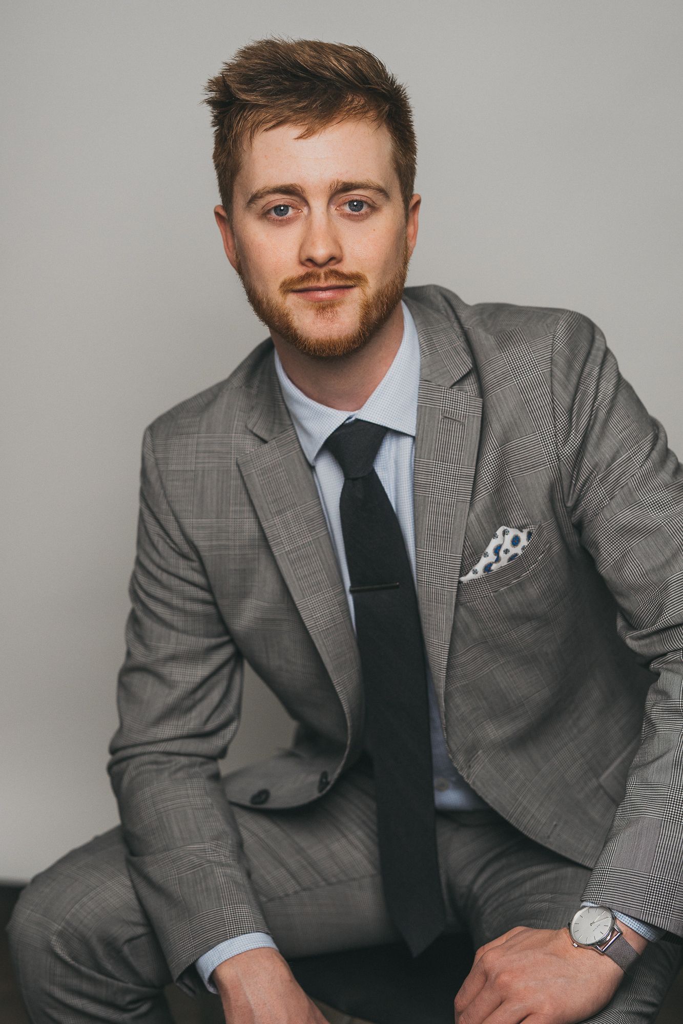 Man in gray plaid suit with black tie and pocket square seated confidently.