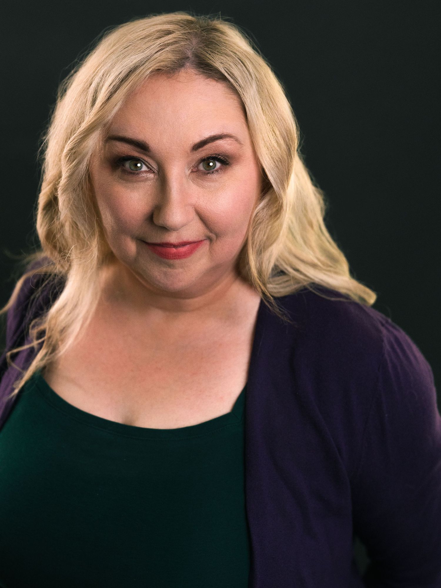 Smiling blonde woman with green eyes wearing a teal top and purple cardigan against a dark background.