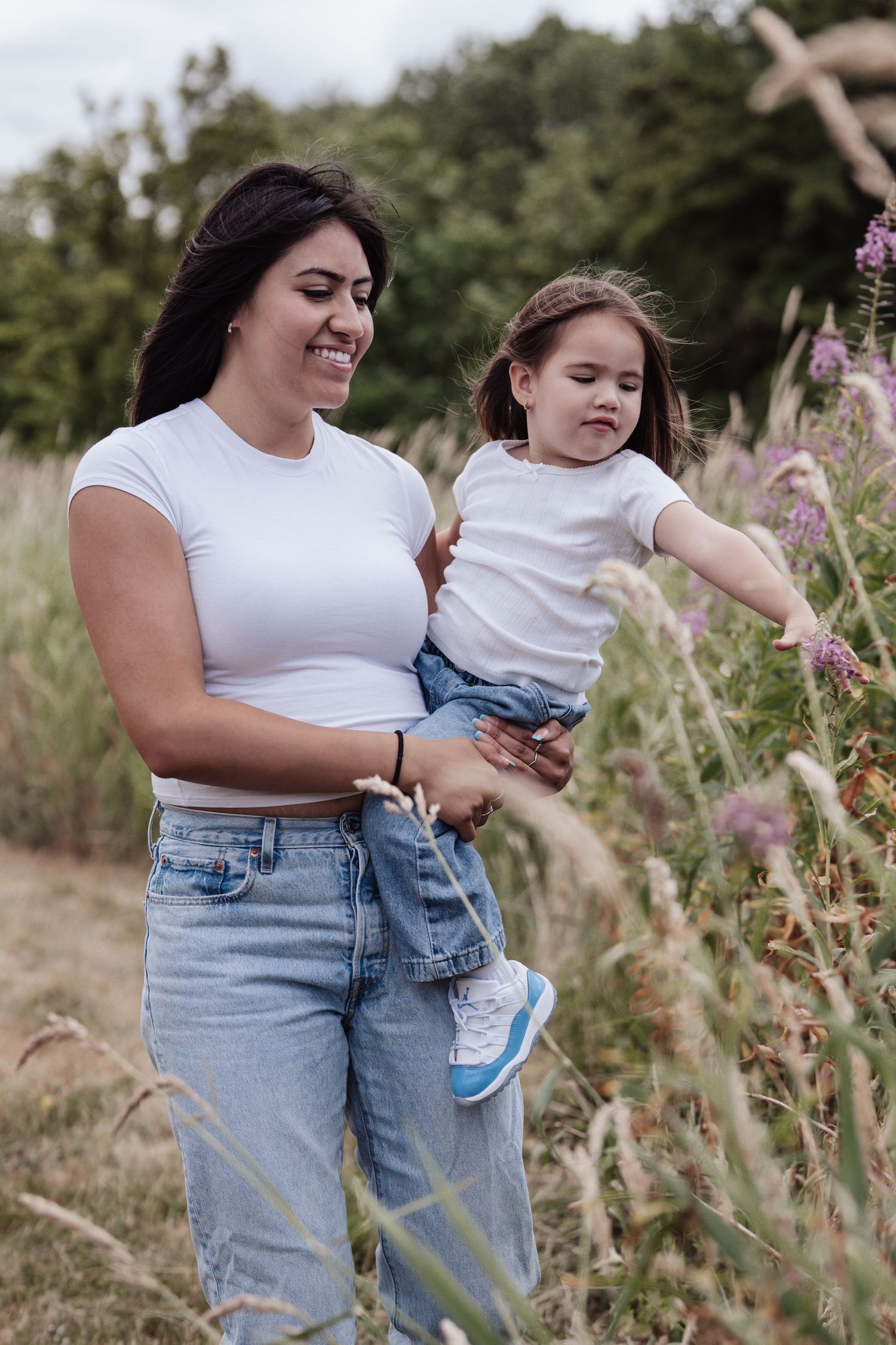 A smiling mother in white top and jeans holds her toddler daughter while exploring wildflowers in a field.