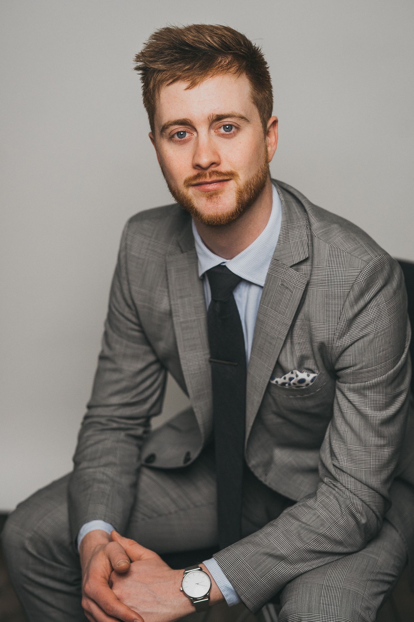 Young man in gray suit with black tie and watch posing with hands clasped.