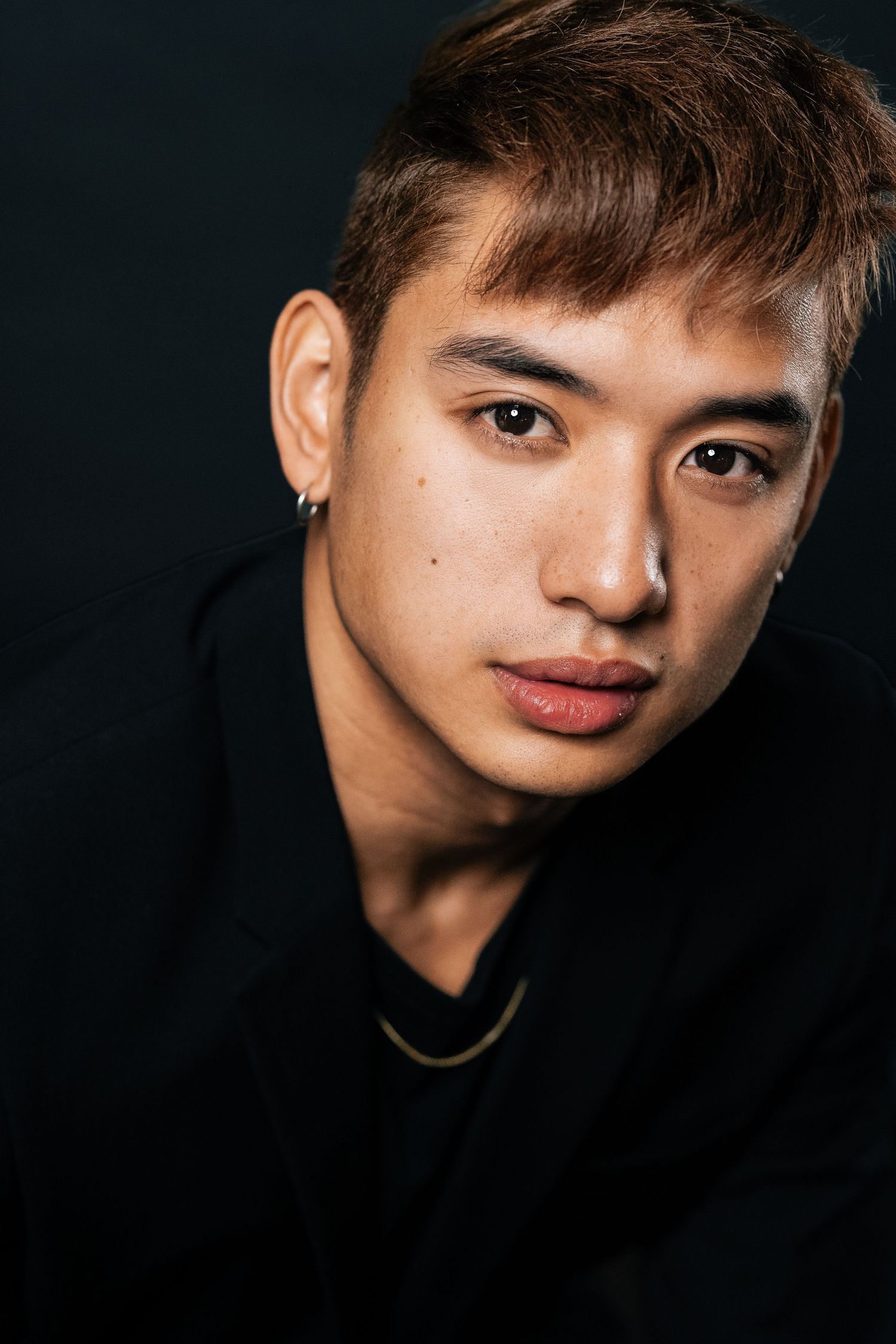 Young Asian man with brown hair, ear stud, and gold chain necklace wearing black jacket against dark background.