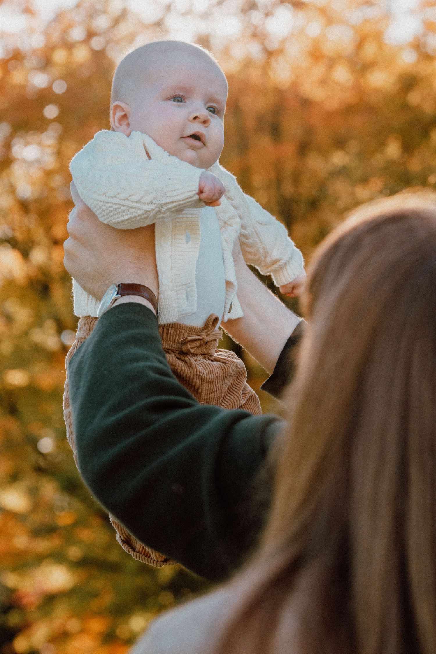 A mother lifts her smiling baby in a white knit outfit against a warm autumn foliage background.