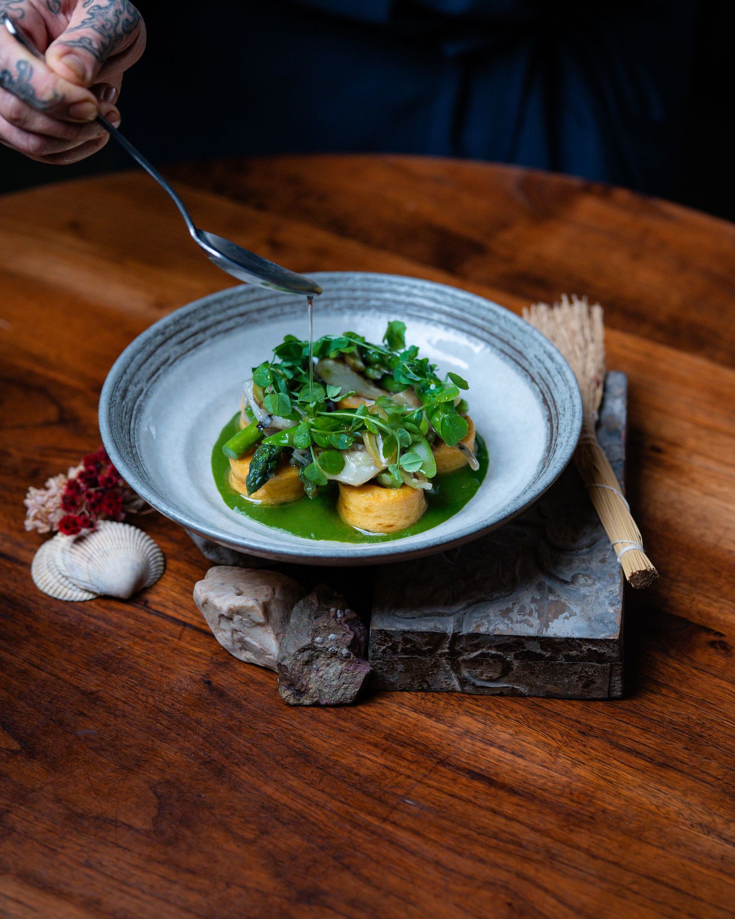 Chef drizzling green herb sauce over gourmet potato and microgreen dish in ceramic bowl on rustic wooden board.