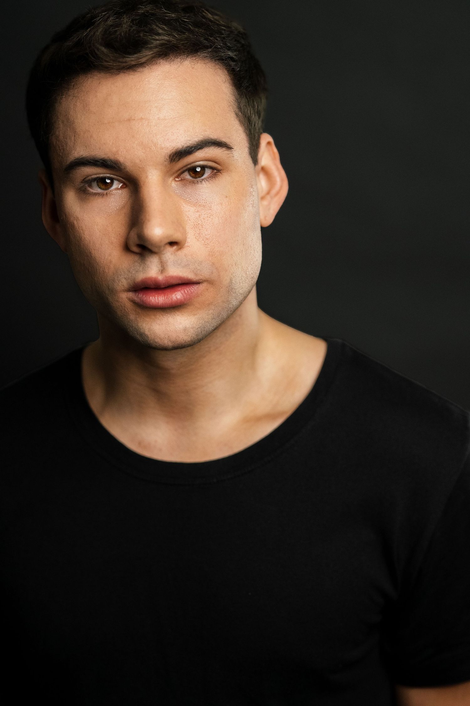 Young man with short dark hair wearing a black t-shirt poses against a dark background in a professional headshot.