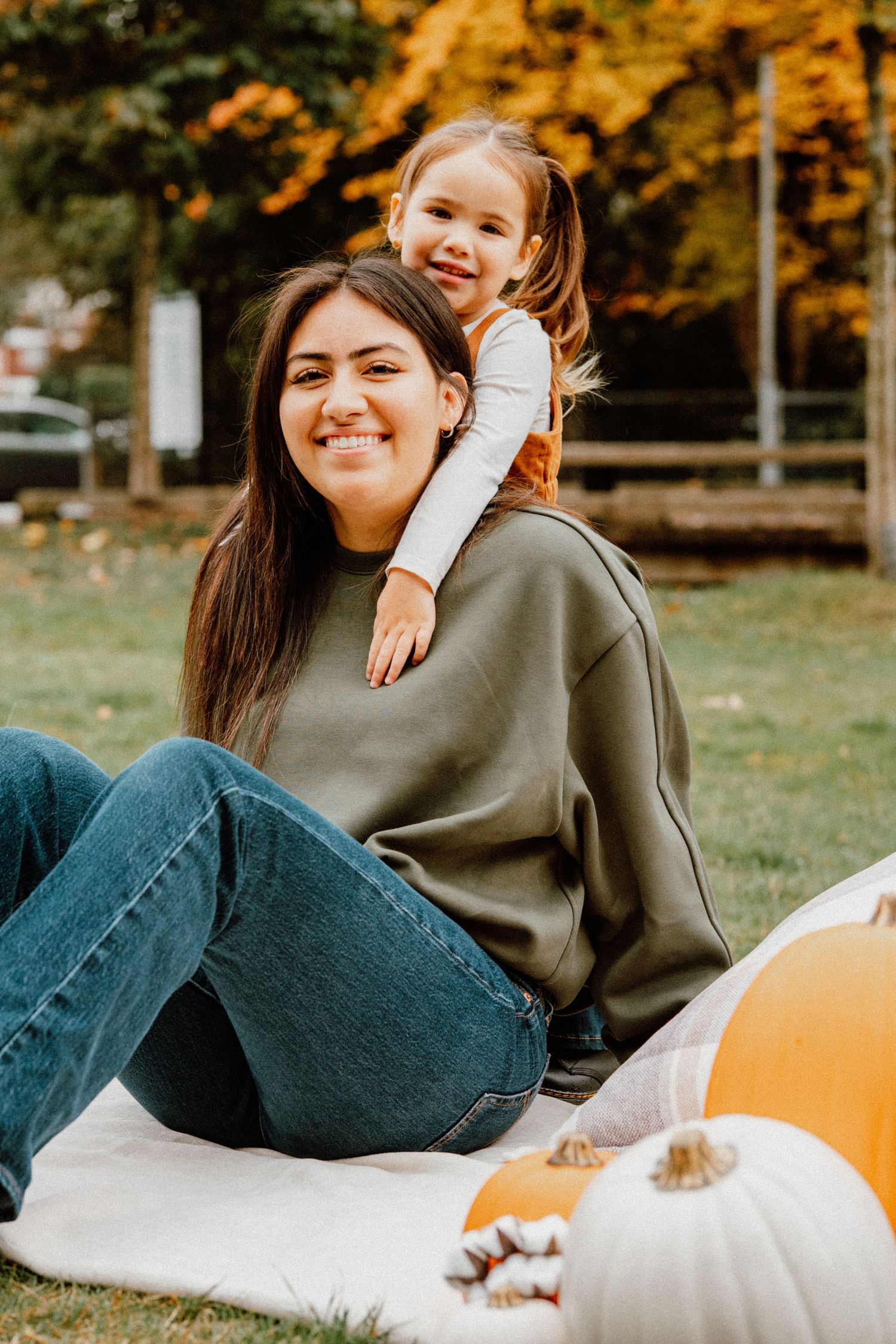 A woman and young girl pose with pumpkins outdoors in autumn, smiling warmly among colorful fall foliage.