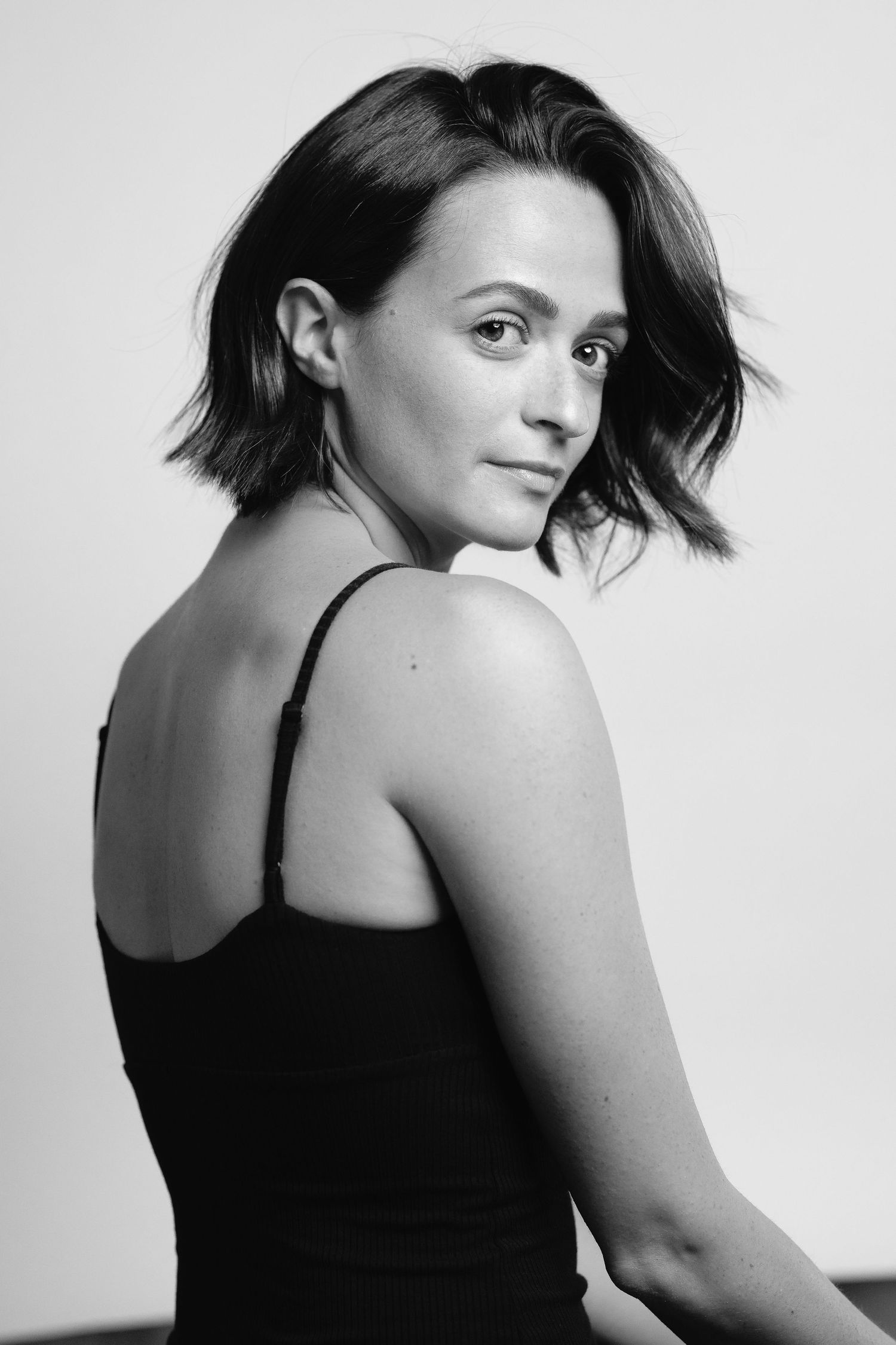 Black and white portrait of a young woman with short dark hair wearing a black tank top, looking over her shoulder.