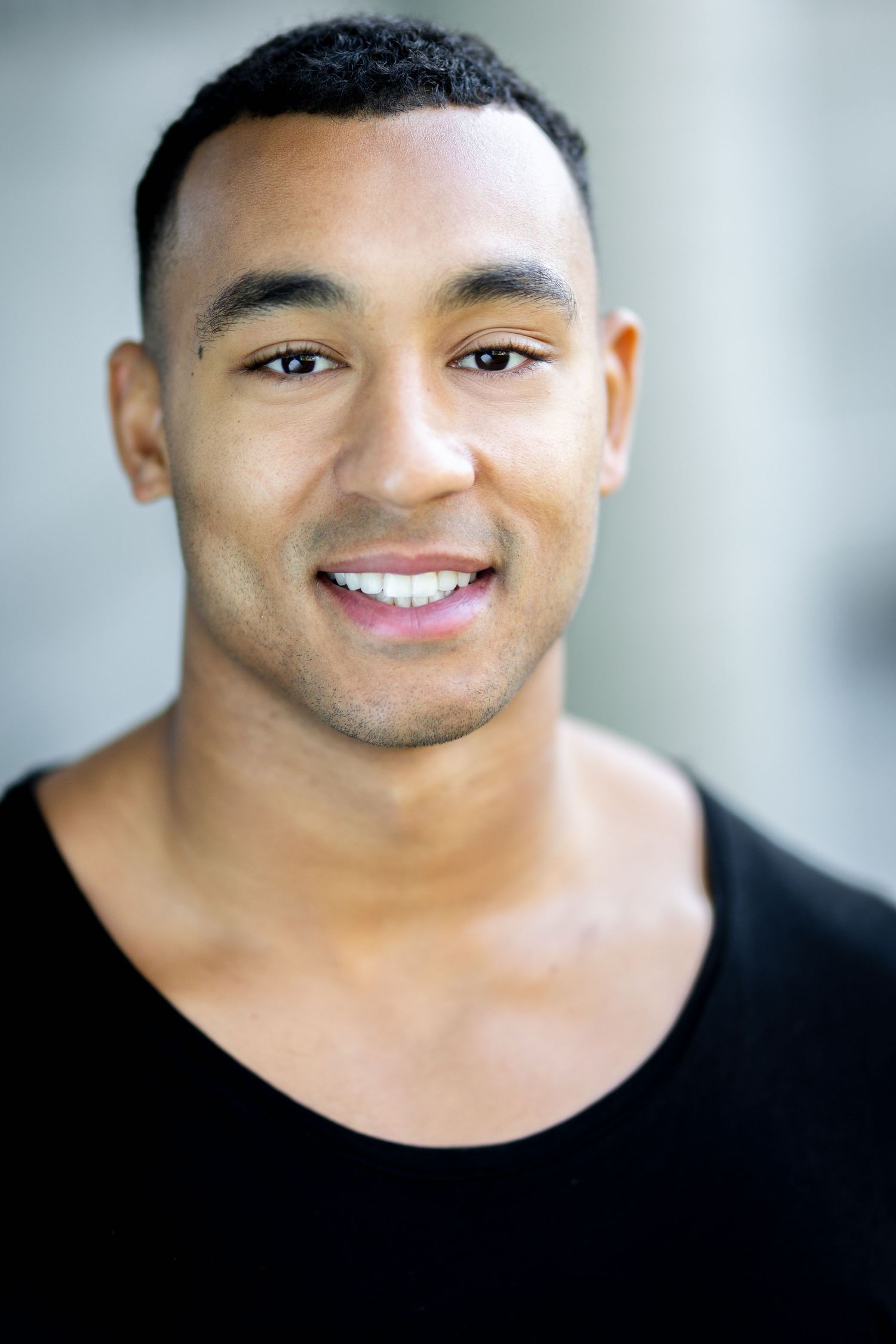 Smiling young man with short dark hair wearing a black t-shirt, posing for a portrait photo outdoors.