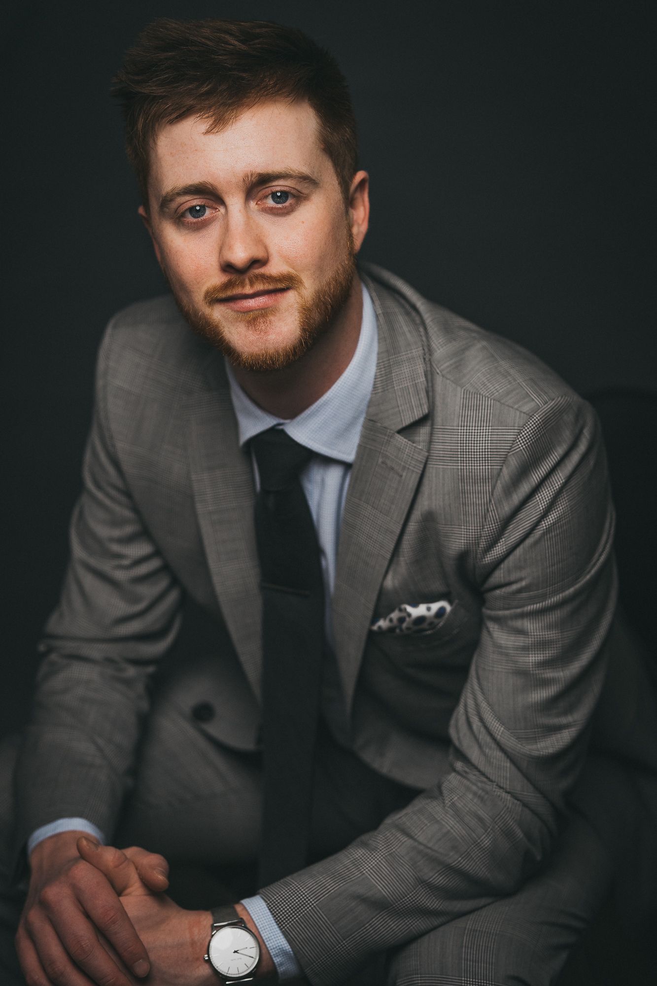 Confident young bearded man in gray plaid suit and black tie posing with hands clasped, wearing a watch.