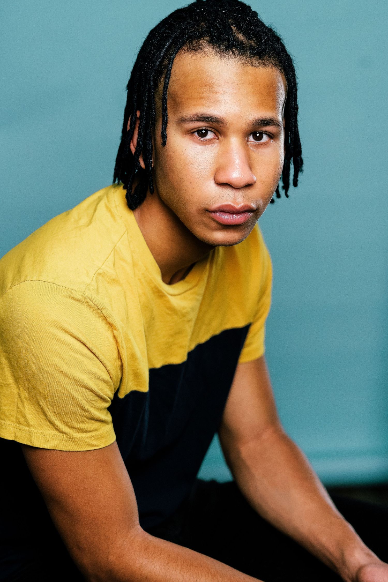 Young man with dreadlocks wearing a yellow and black t-shirt poses against a teal background.