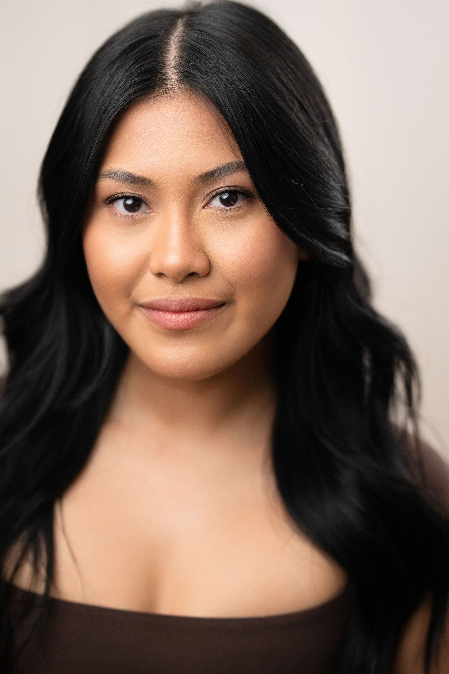 Young woman with long black wavy hair smiling softly, wearing a brown top against a neutral background.