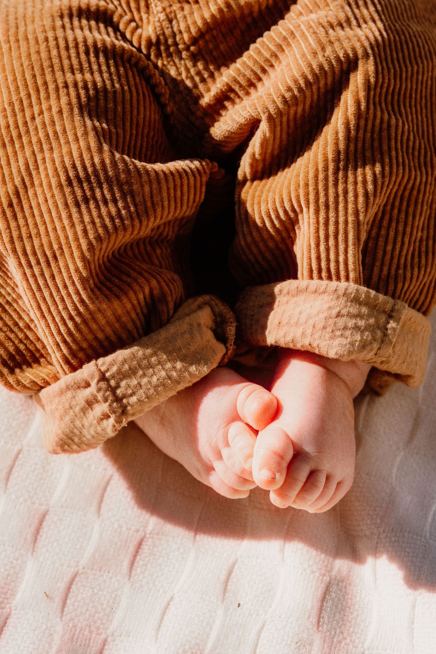 Close-up of tiny baby feet in brown corduroy pants resting on a white knit blanket in warm sunlight.