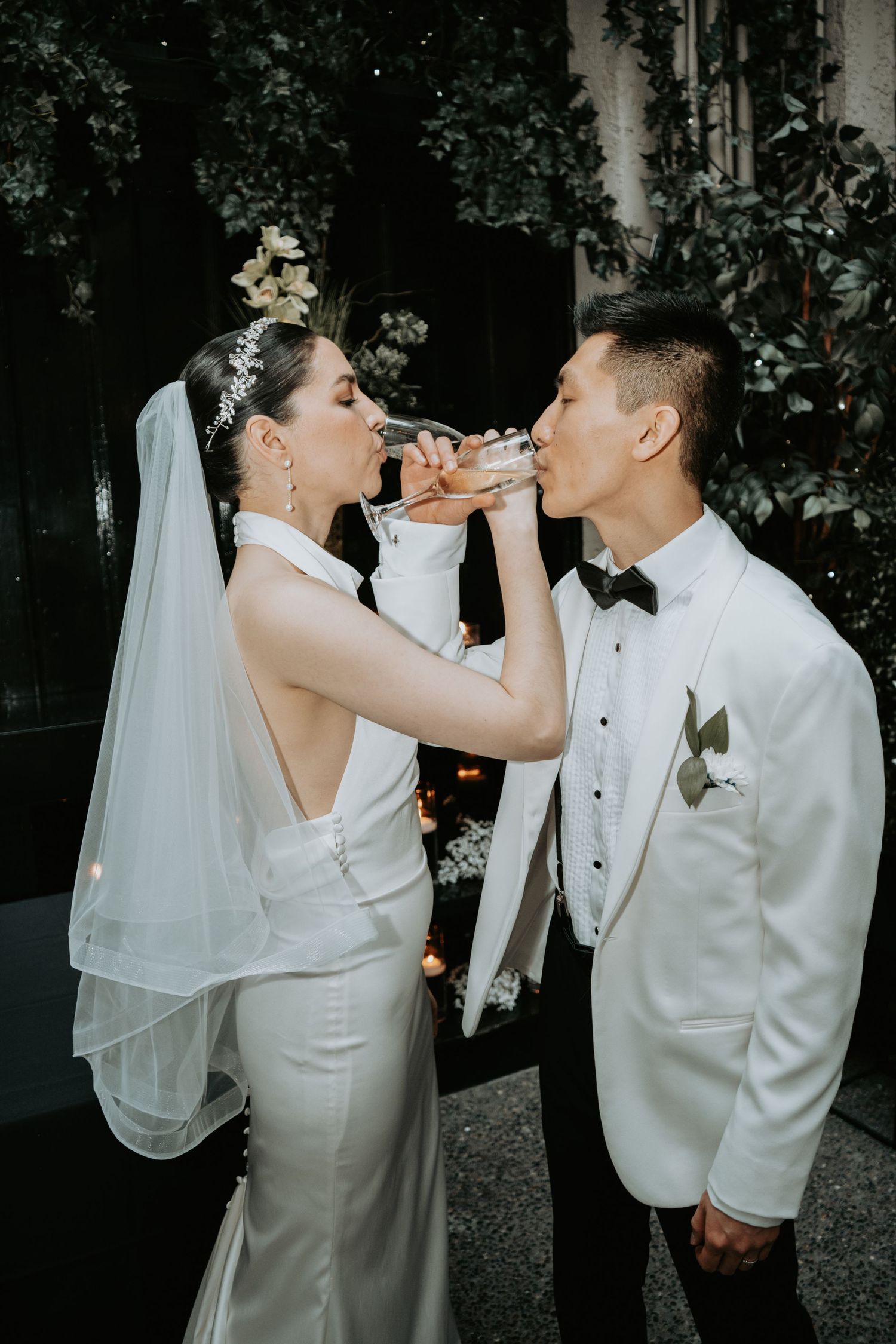 Bride and groom in white attire crossing arms to drink champagne together at elegant wedding celebration.