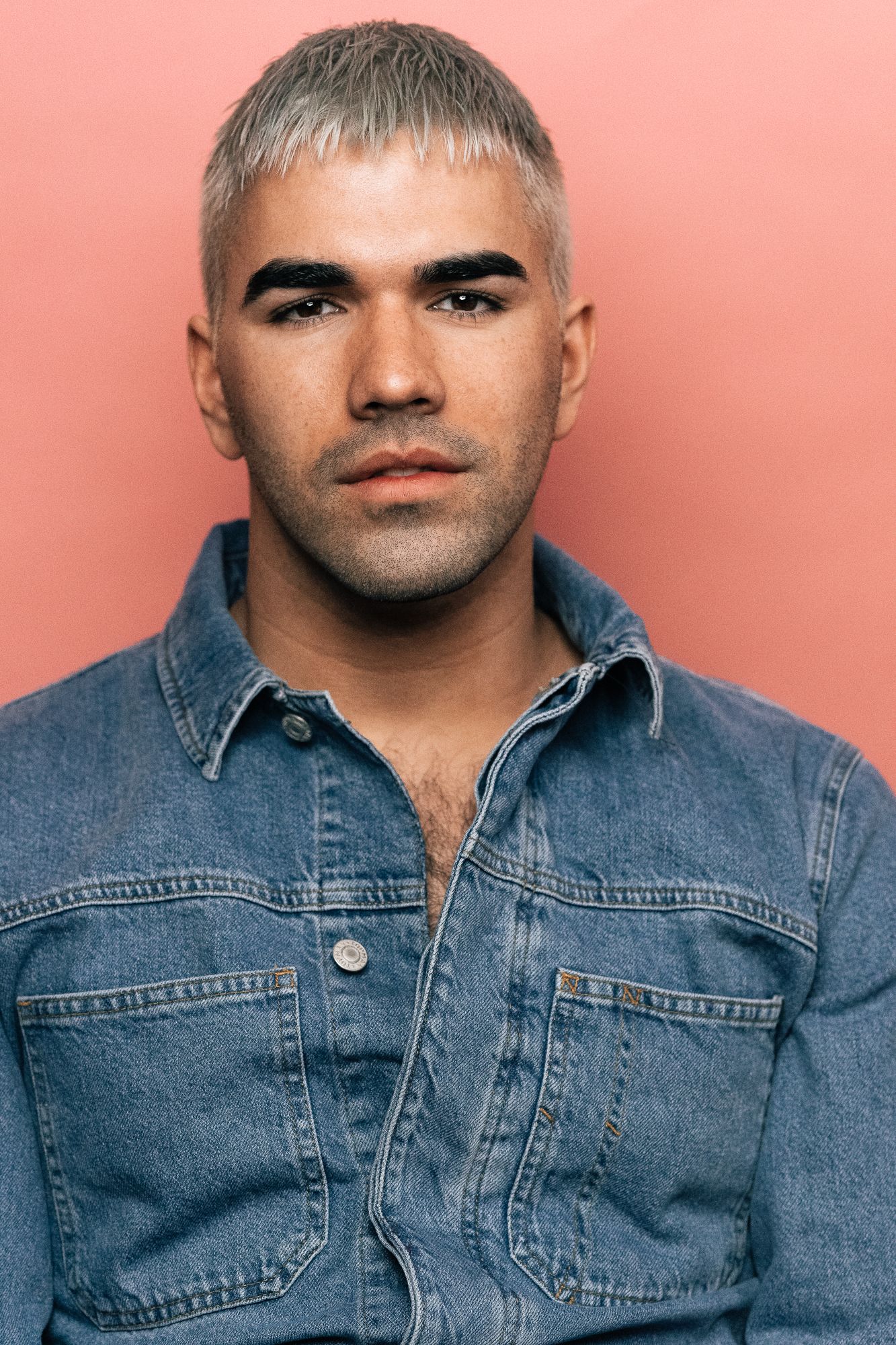 Young man with bleached hair and dark brows wearing an open denim shirt, posing against a pink background.