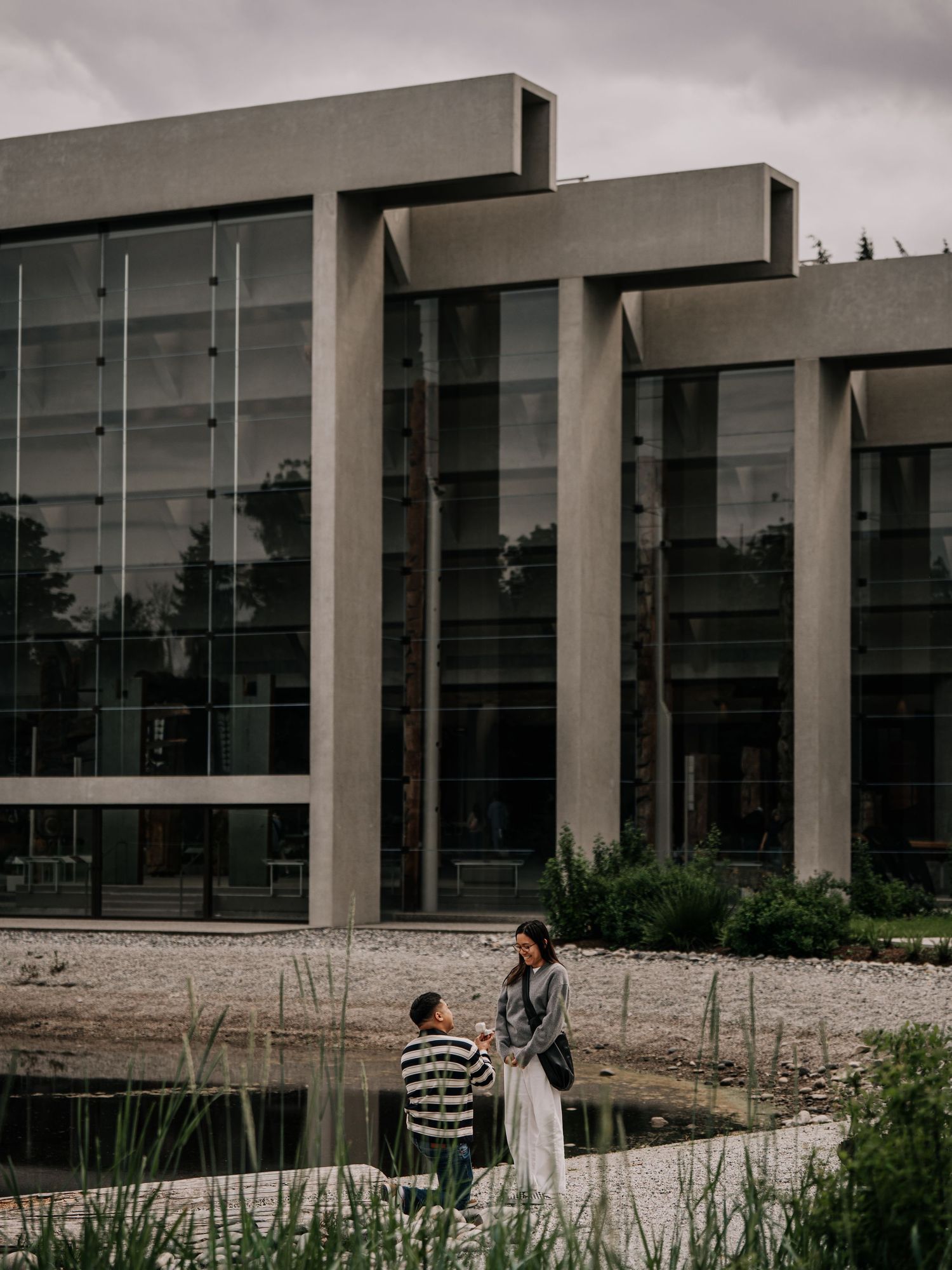 Couple standing by a reflective pond in front of a modern brutalist glass and concrete building on a cloudy day.