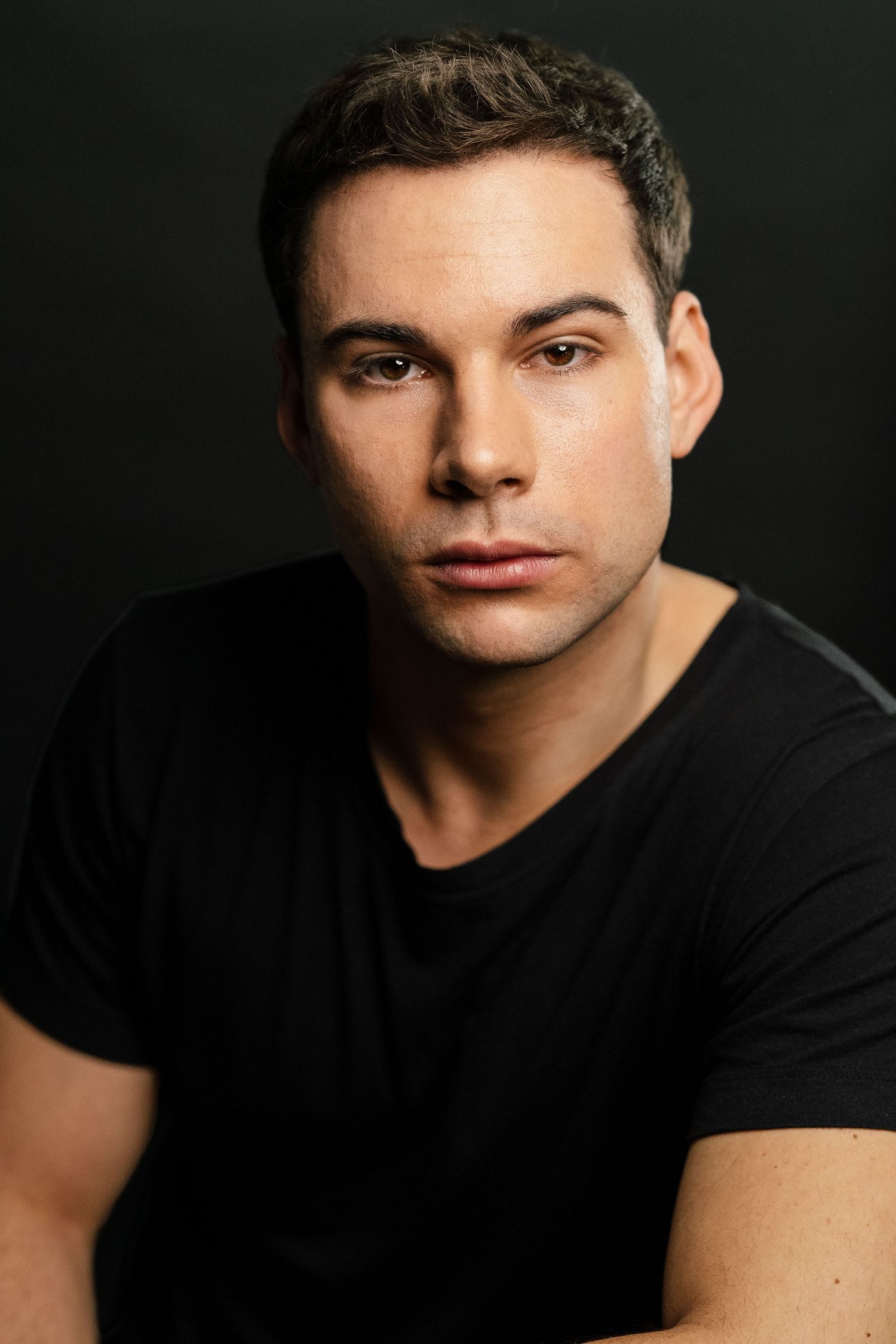 Young man with short dark hair wearing a black t-shirt poses against a dark background in a professional headshot.