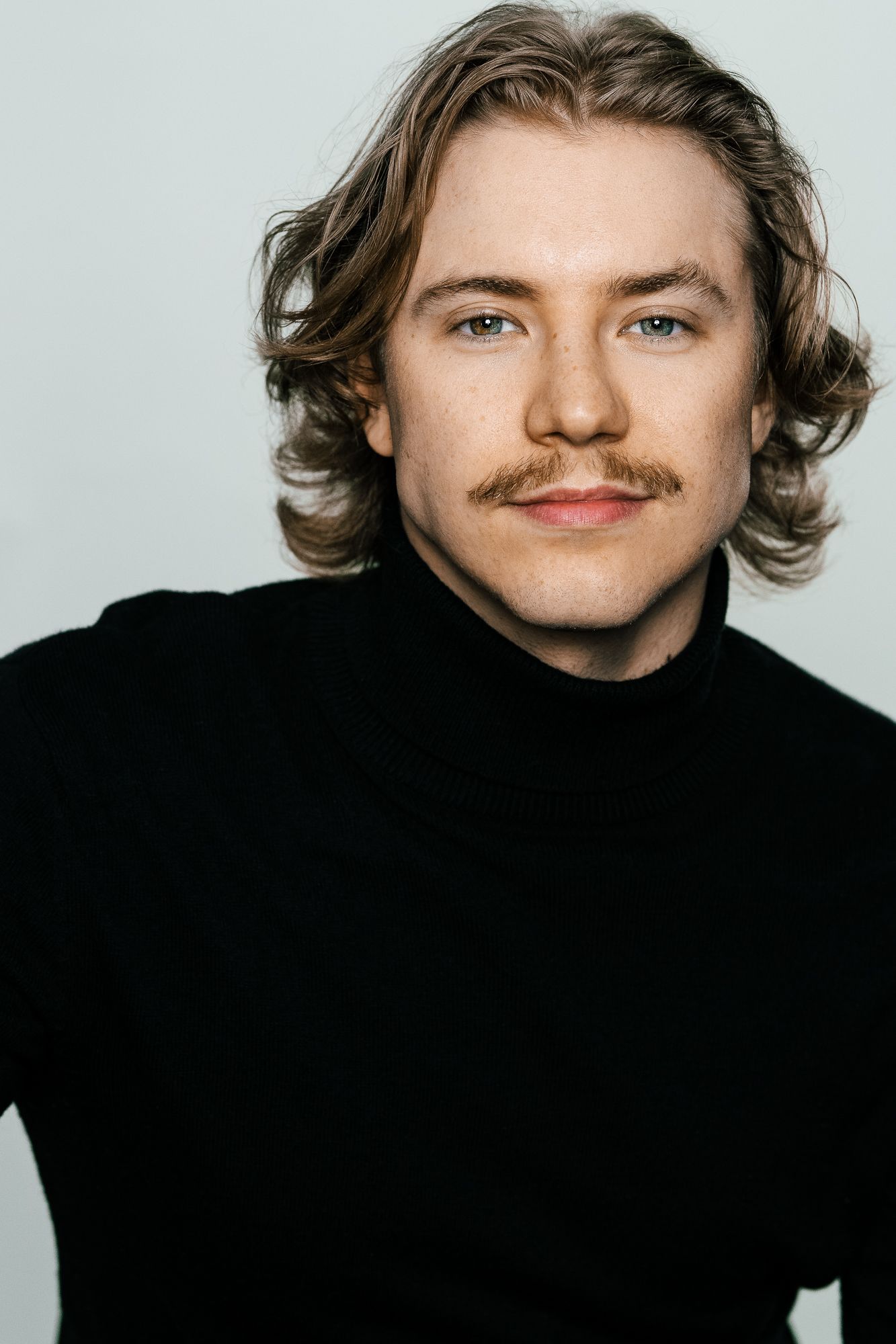 Young man with wavy blonde hair and mustache wearing a black turtleneck sweater against a light gray background.