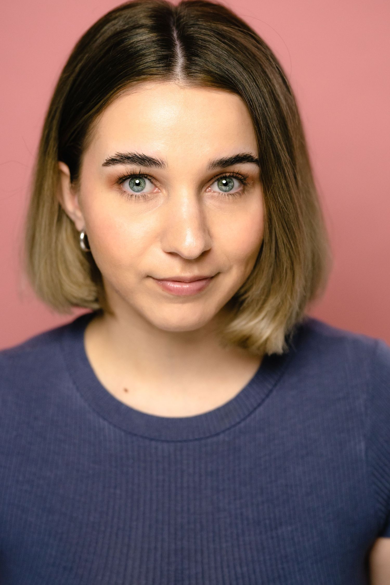 Young woman with blue eyes and blonde bob haircut wearing navy blue top against pink background.