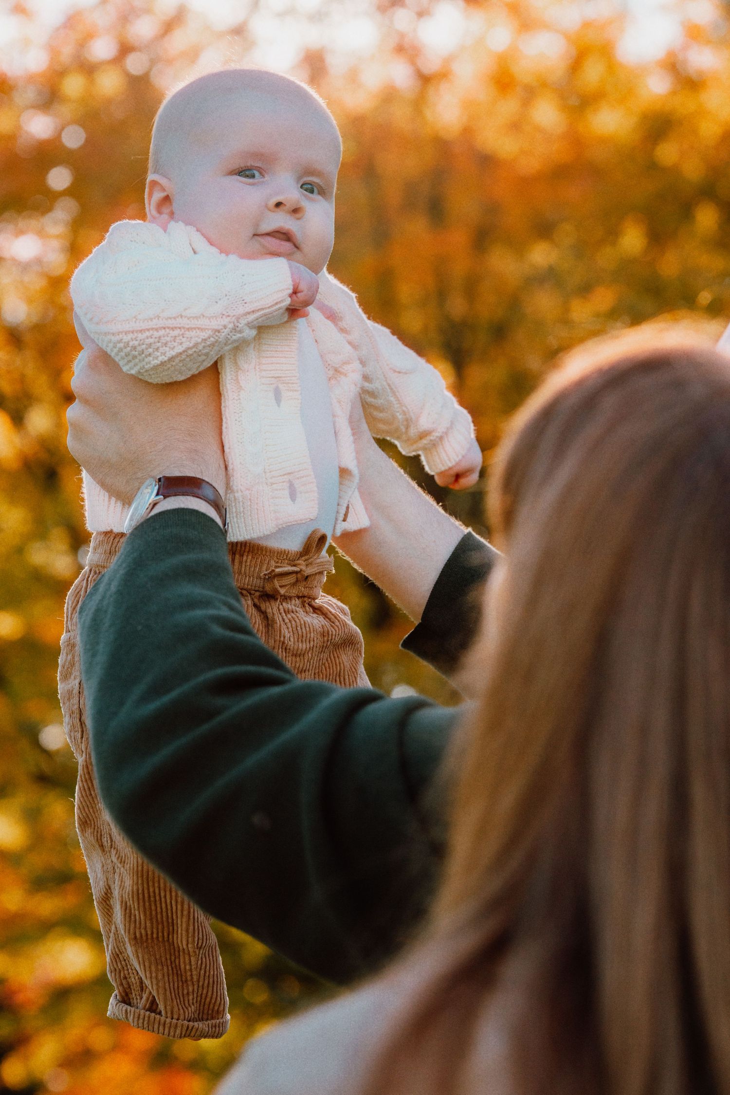 A mother holds her baby up in the air outdoors during autumn, surrounded by golden fall foliage.