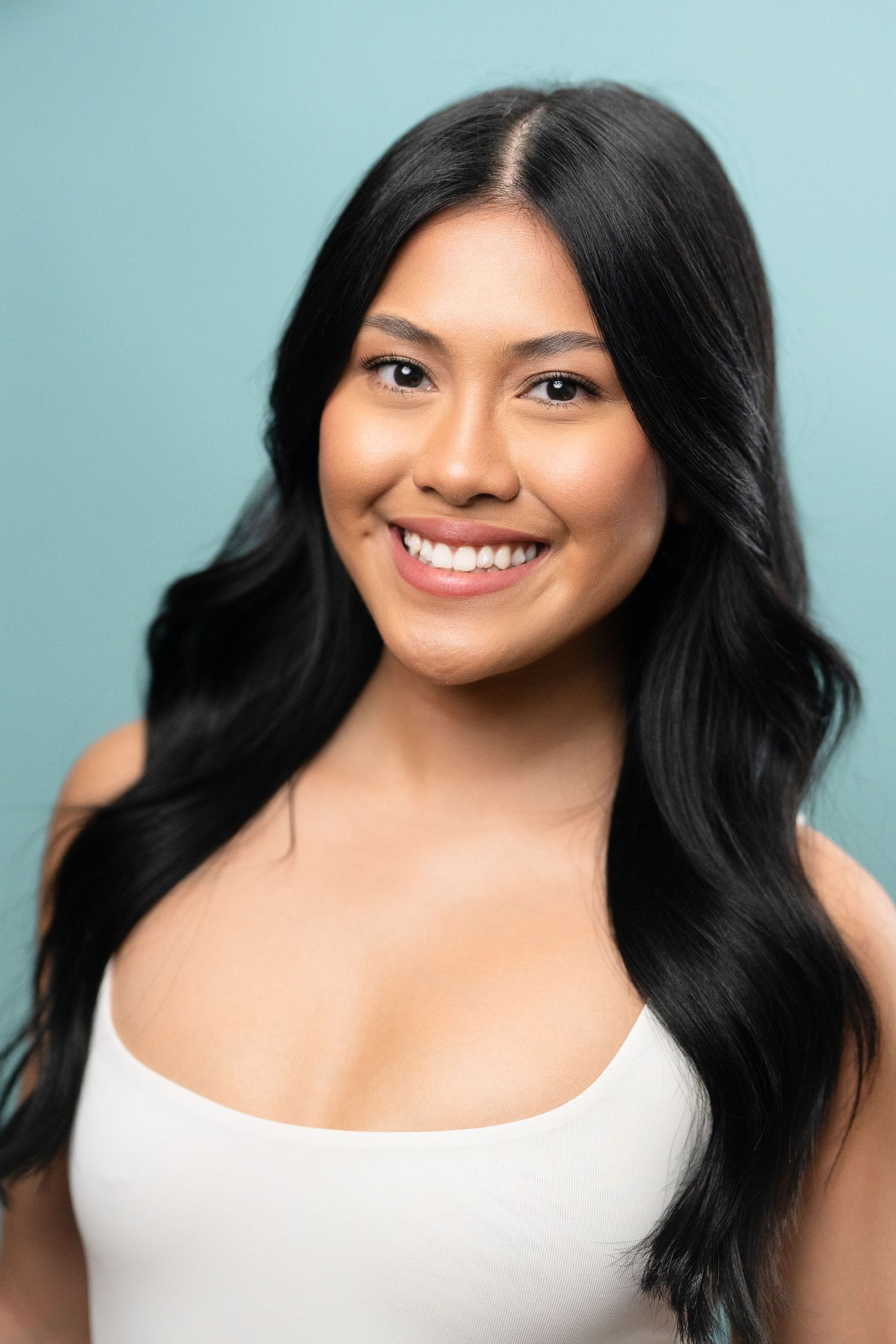 Smiling young woman with long black wavy hair wearing a white tank top against a light blue background.