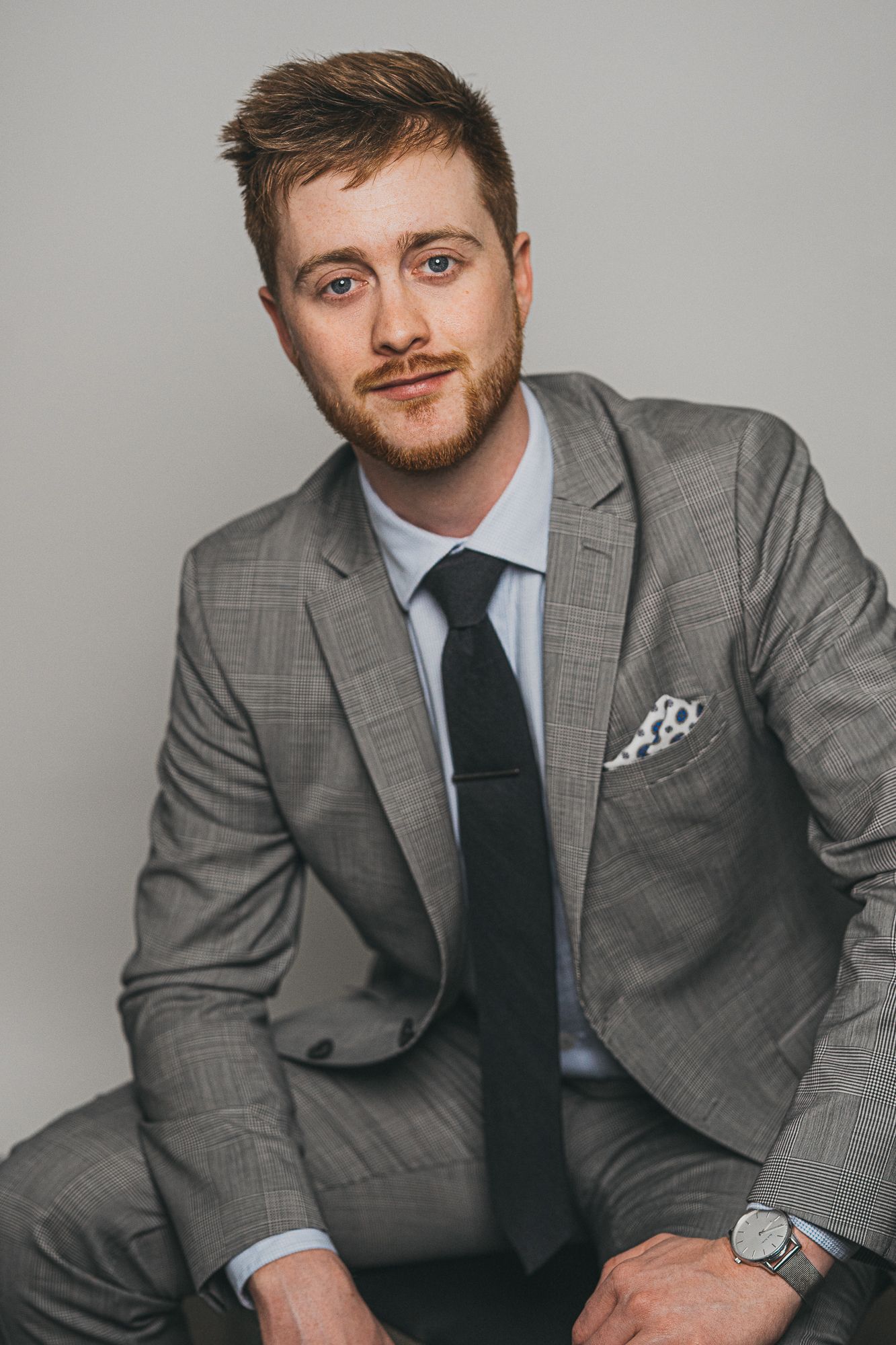 Young man with red beard wearing a gray plaid suit, black tie, and white pocket square, posing confidently.