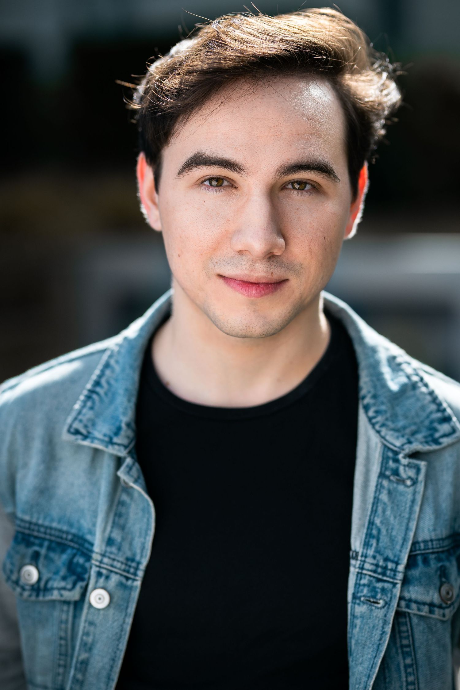 Young man in denim jacket and black shirt smiling confidently outdoors in a professional headshot photo.