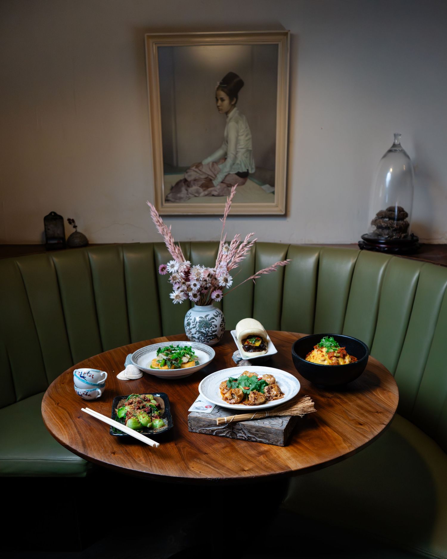 Round wooden table with Asian dishes, green booth seating, floral vase, and portrait painting in a cozy restaurant setting.