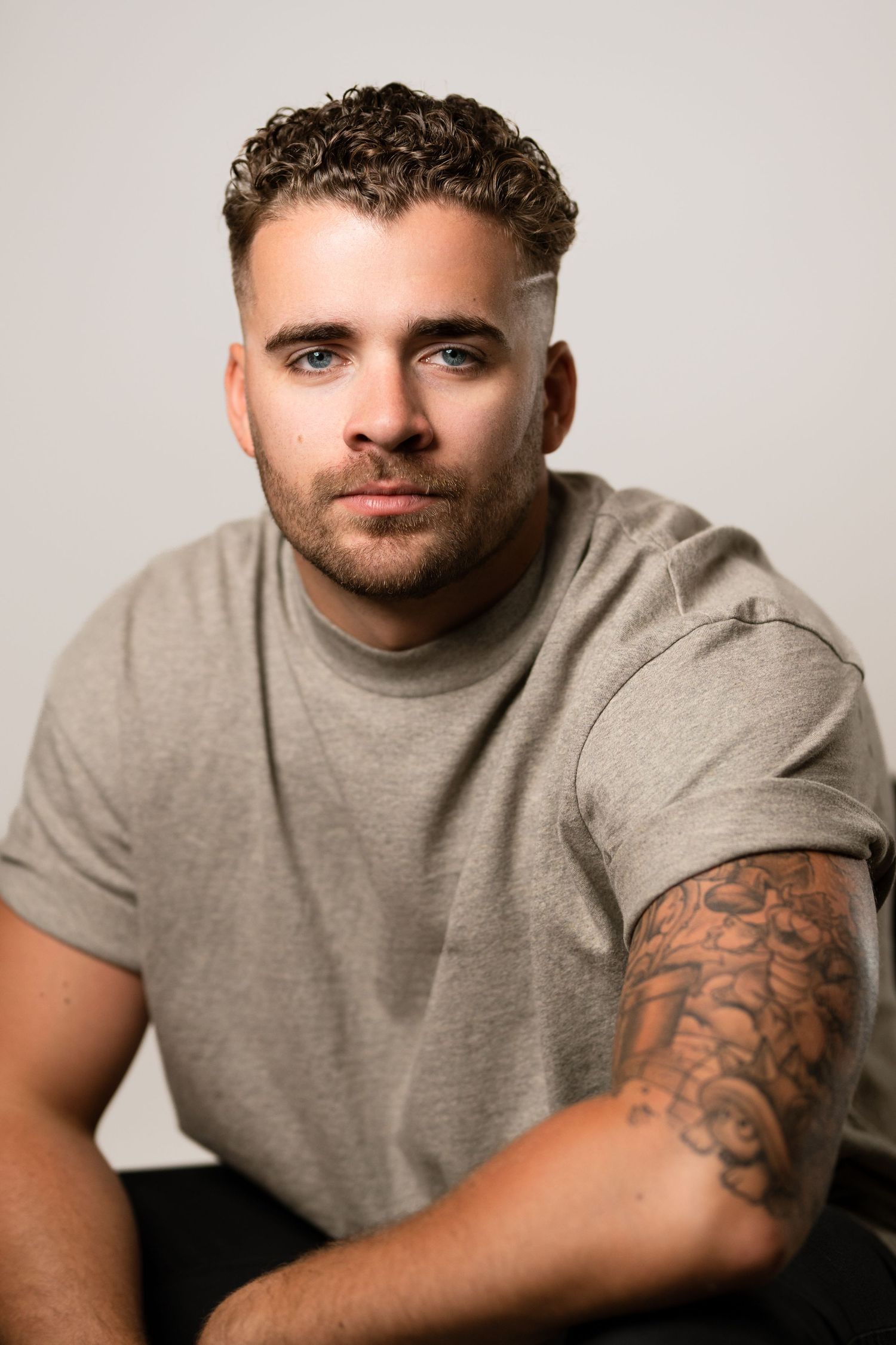 Young man with curly hair, blue eyes, and arm tattoos wearing a gray t-shirt poses for a studio portrait. Headshots Vancouver