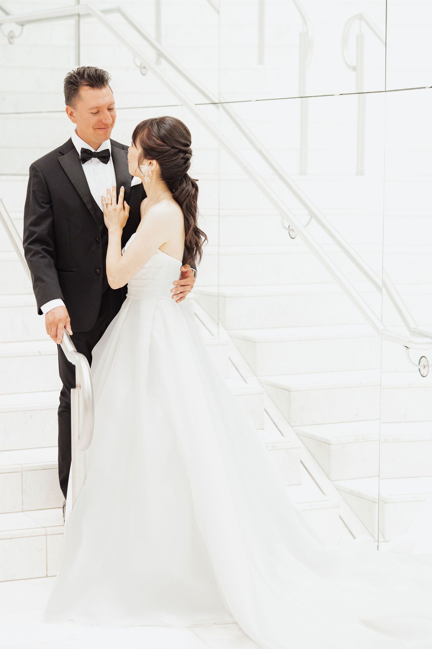 Bride in white ball gown and groom in black tuxedo share a romantic moment on elegant white staircase.