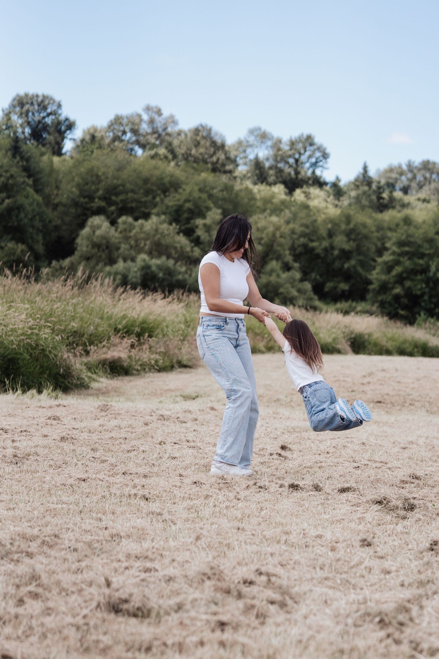 A mother swings her young daughter by the hands in an open field with lush green trees in the background.