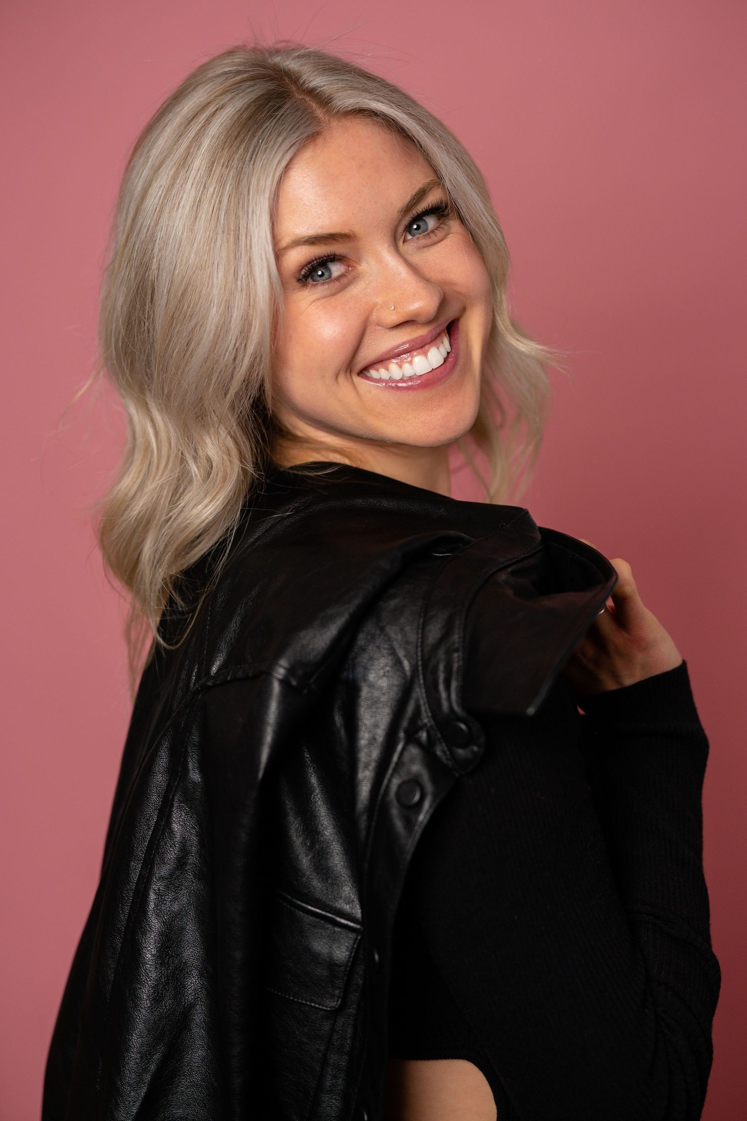 Smiling blonde woman in black leather jacket poses against a pink background.