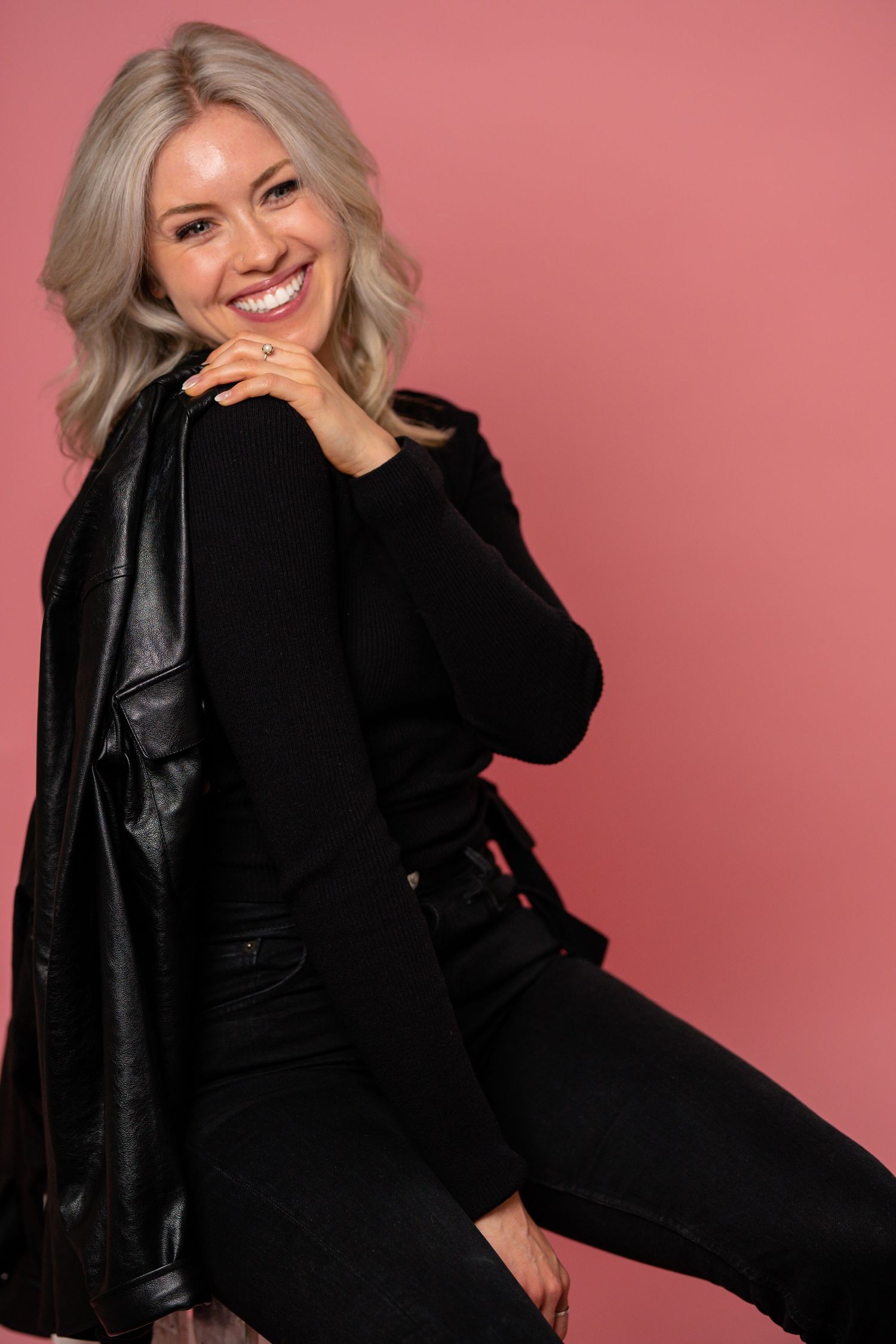 Smiling blonde woman in all-black outfit with leather jacket poses against a pink background.