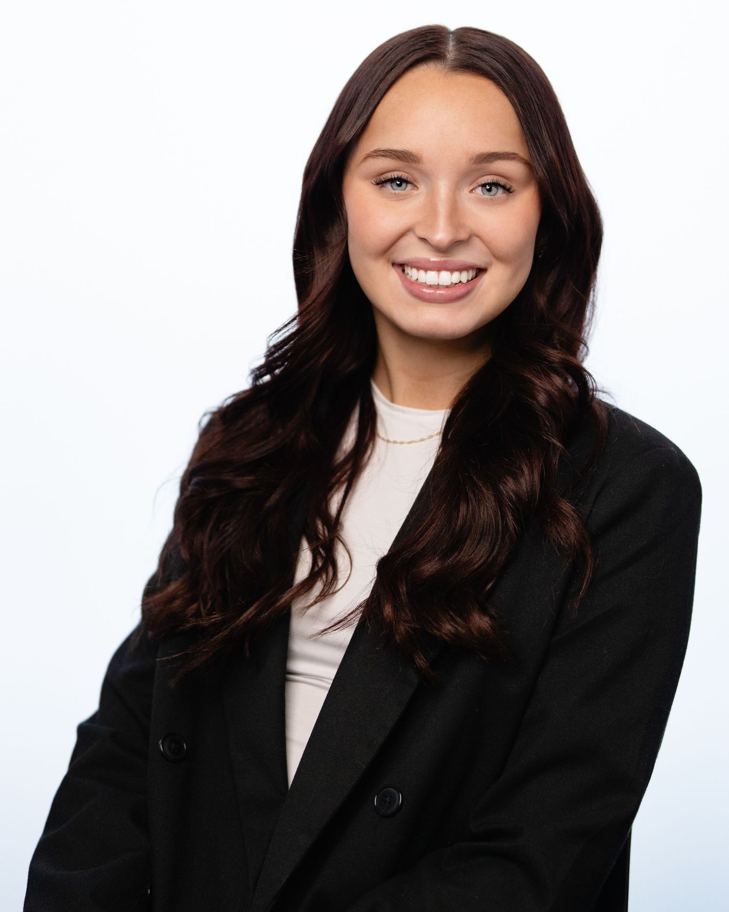 Young professional woman with long dark wavy hair smiling in a black blazer against a white background. Professional Headshots Vancouver